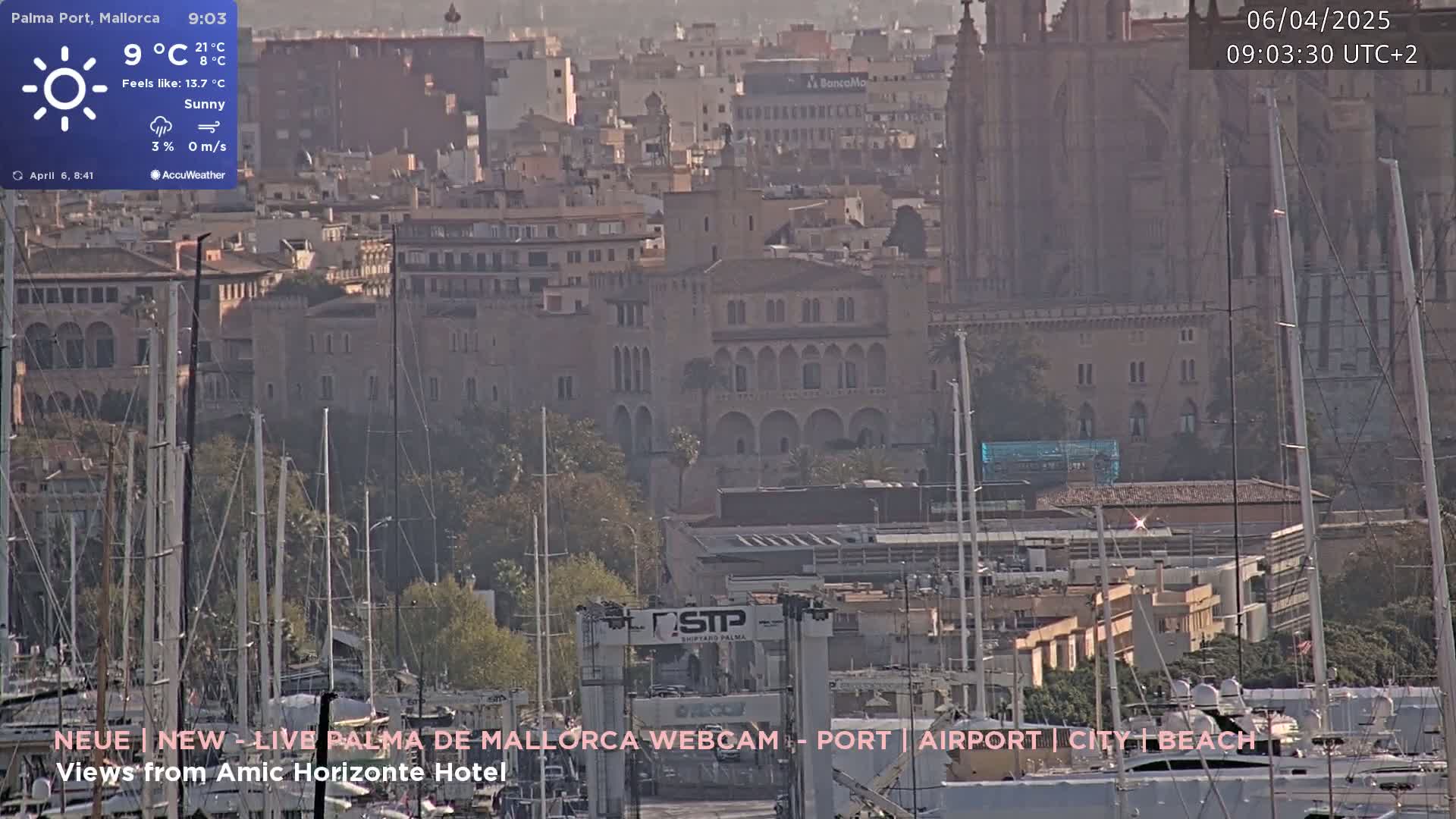 A sunny harbor scene shows numerous masts of sailboats in the foreground, with a city and a large cathedral in the background.