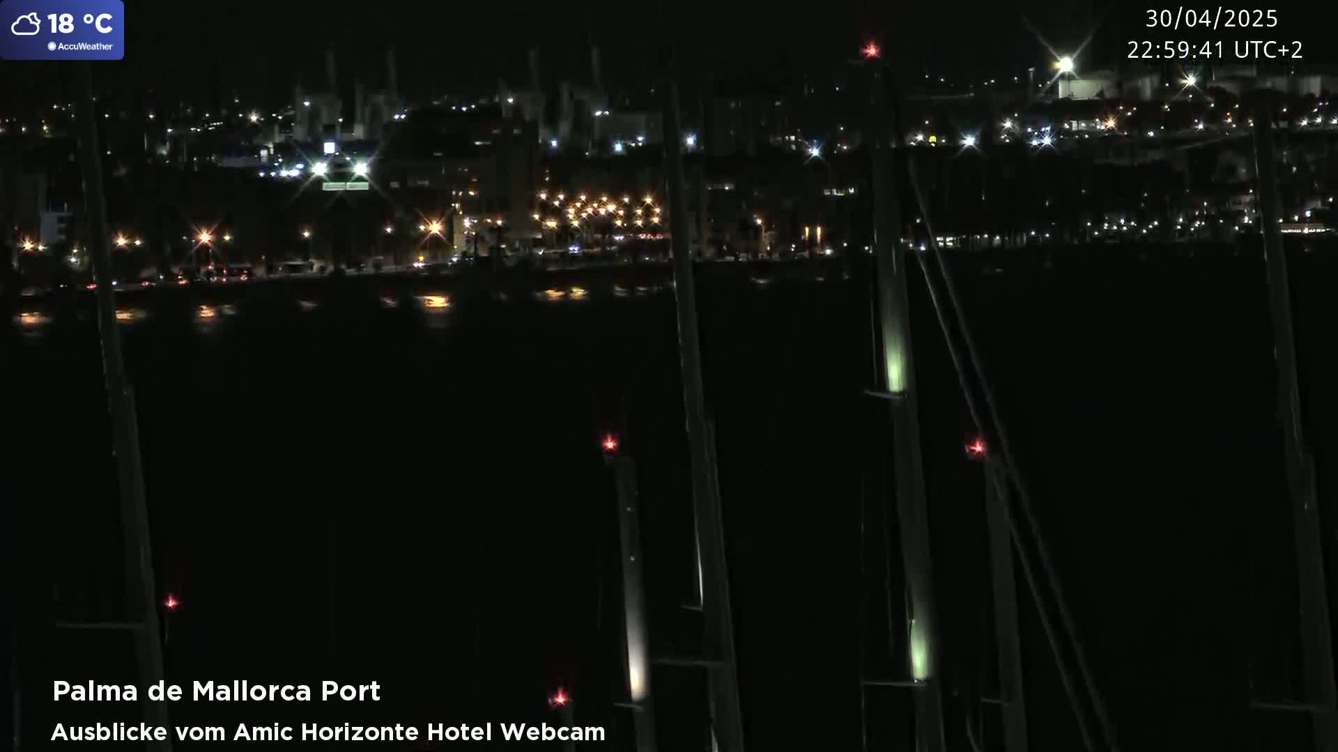 A nighttime view of a city harbor with lights illuminating the waterfront and boats, under a clear sky with a temperature of 18 degrees Celsius.