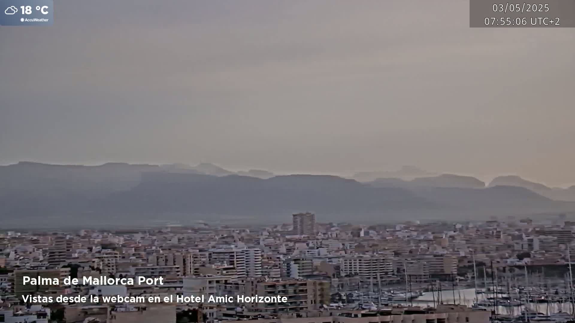 A hazy, 18°C day reveals a cityscape with a harbor full of boats, backed by distant mountains.