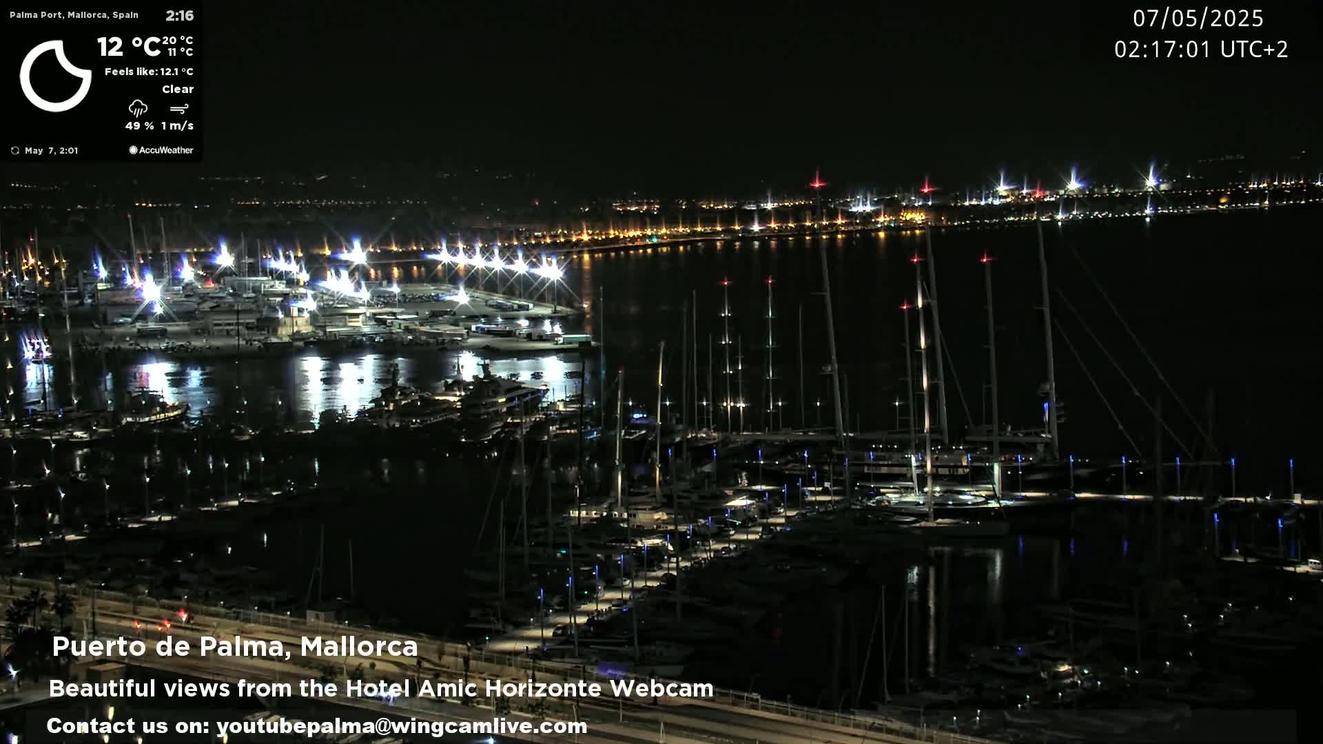 A nighttime, clear view of a marina filled with boats, illuminated by lights, with a city skyline in the distance.