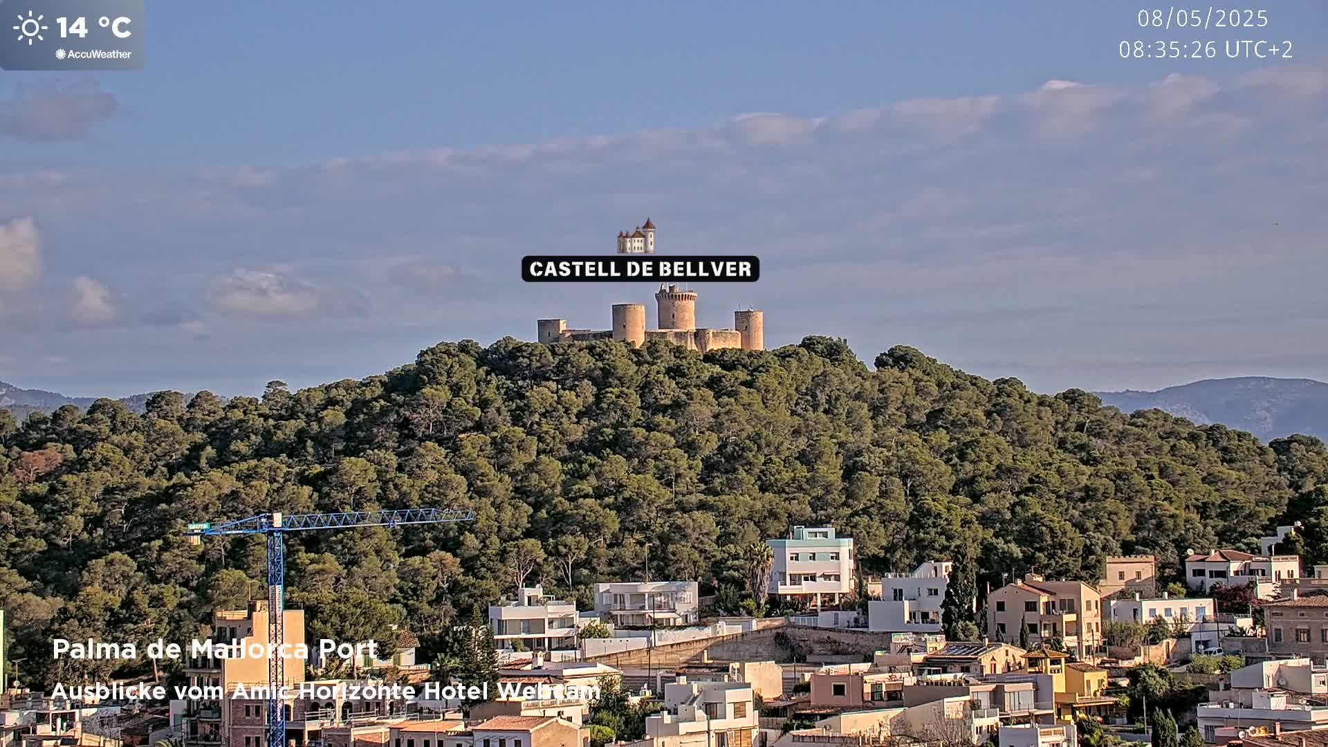 A stone castle sits atop a tree-covered hill overlooking a town under a partly cloudy sky.