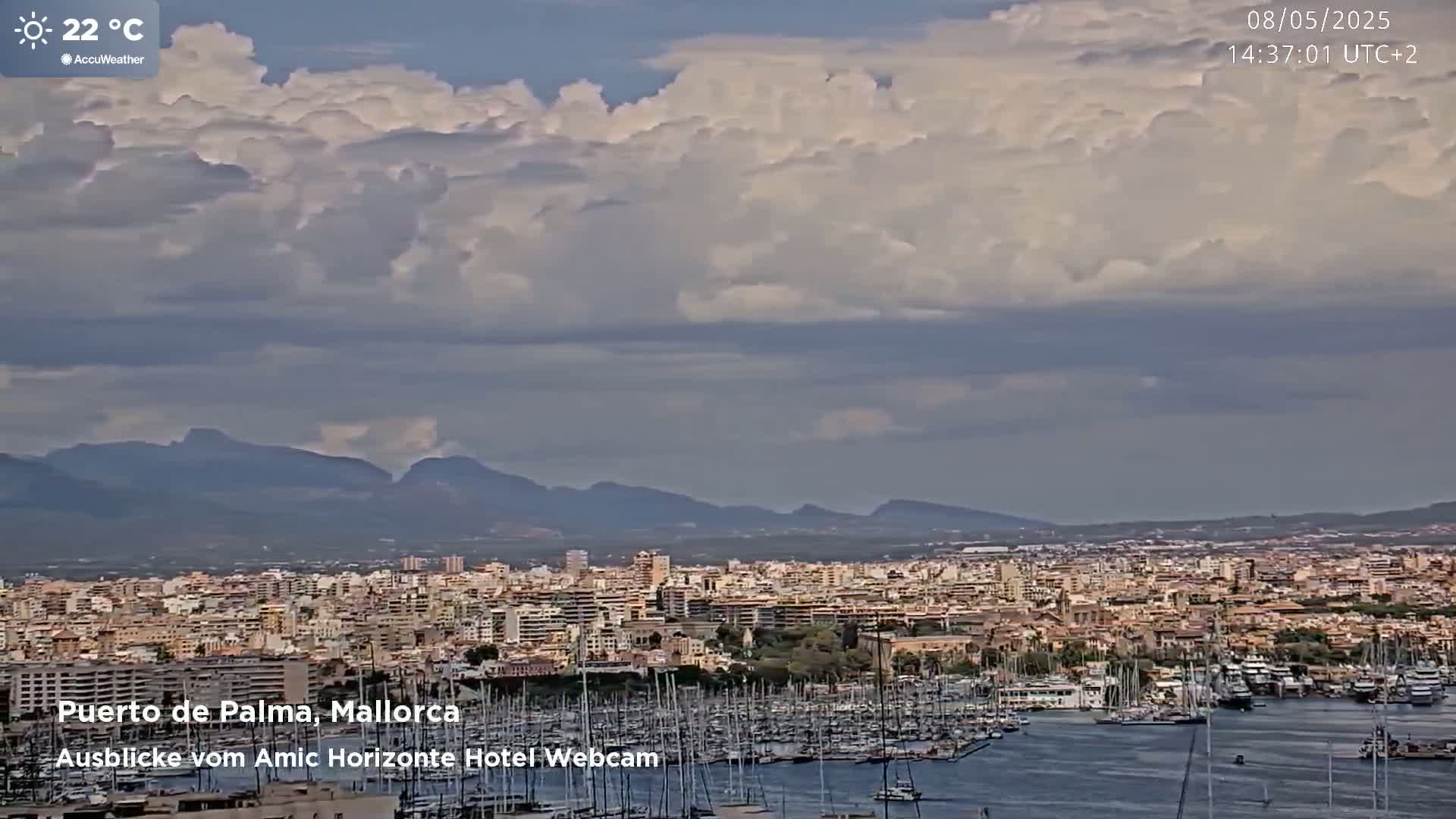 A partly cloudy sky overlooks a harbor filled with numerous boats and a sprawling city with mountains in the distance.