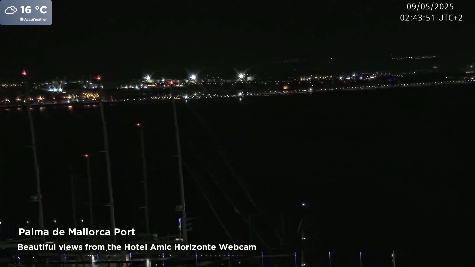A nighttime view of a harbor with many masts and a city skyline illuminated in the distance under a partly cloudy sky.