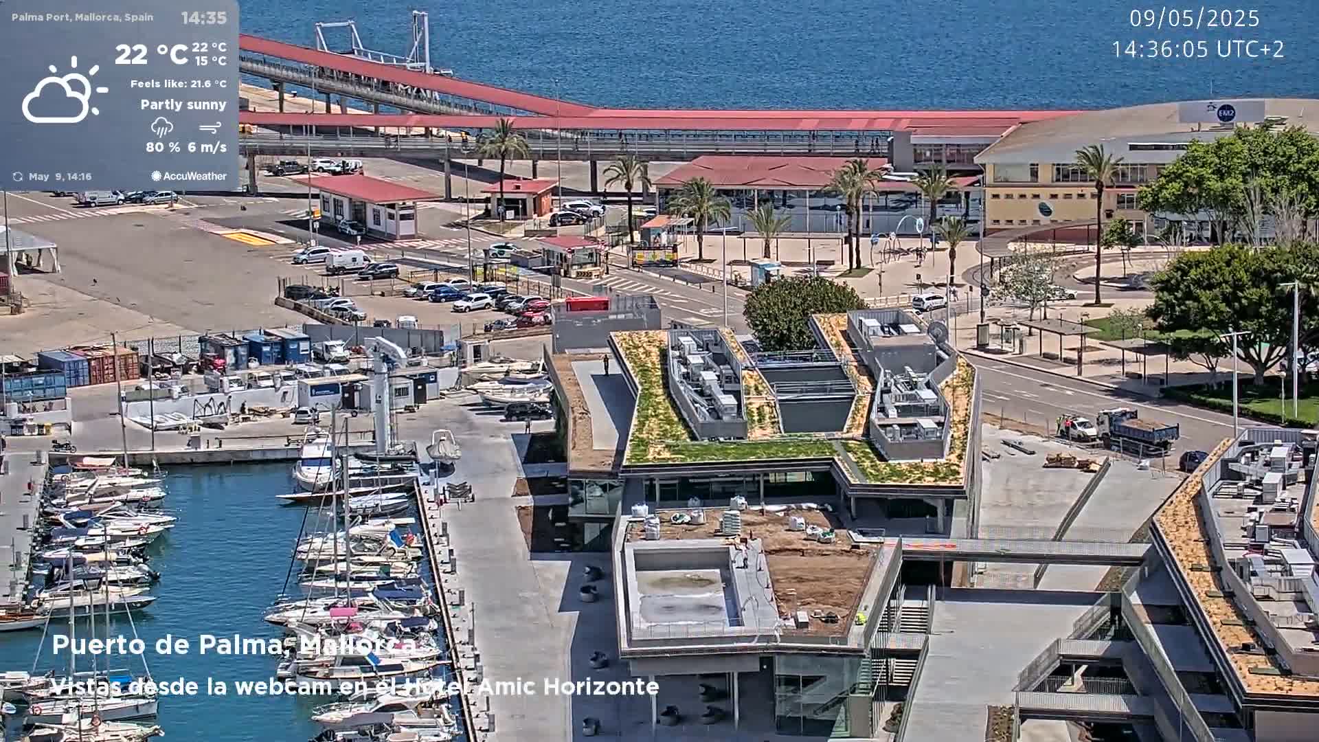 A harbor marina with numerous boats, partially sunny weather, and a modern building with a green roof under construction is visible from an elevated vantage point.