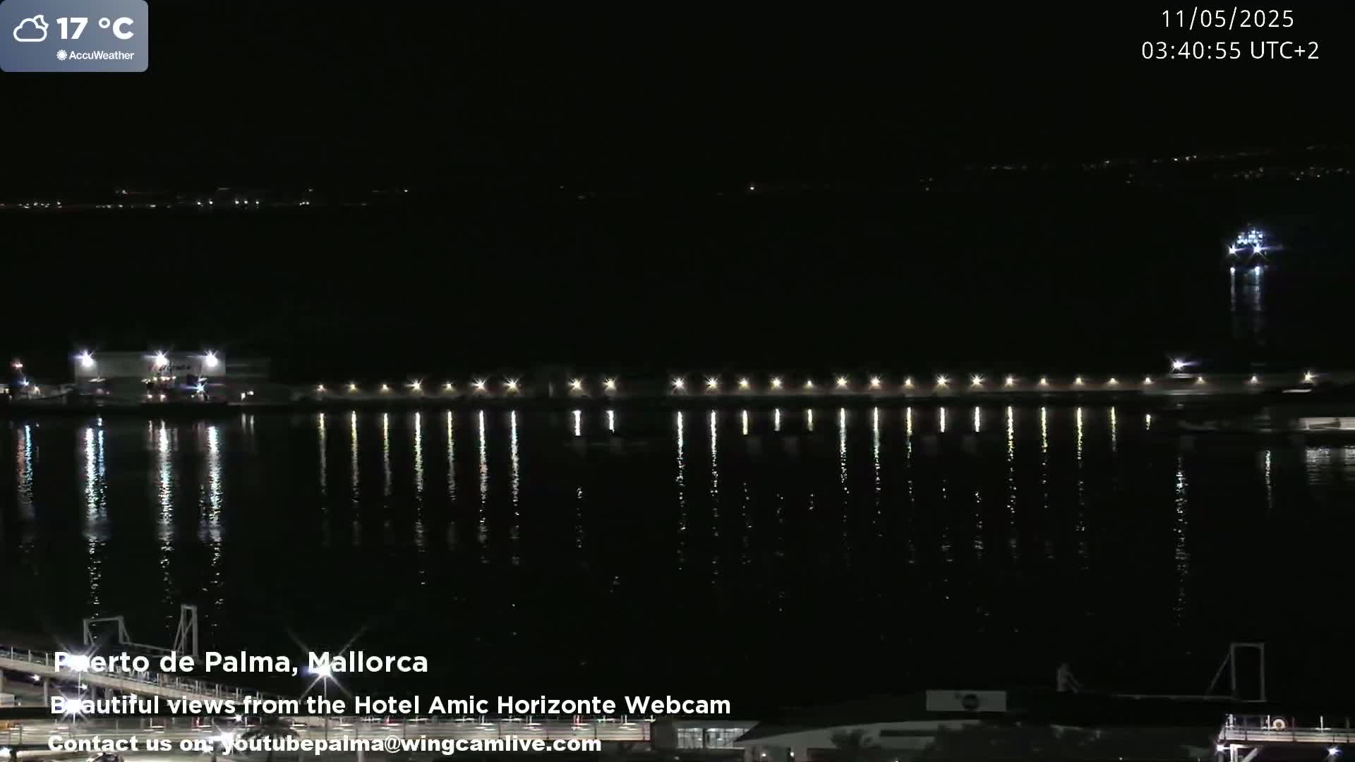 A nighttime harbor scene shows a calm body of water reflecting the lights of a pier and distant shoreline under a cloudy sky.