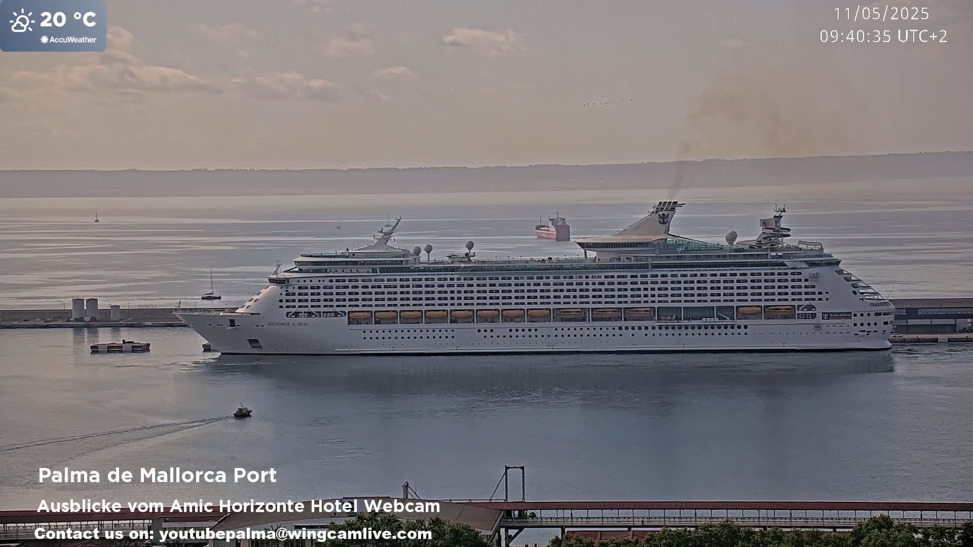 A large cruise ship is docked at a port on a hazy, partly cloudy day with a temperature of 20°C.
