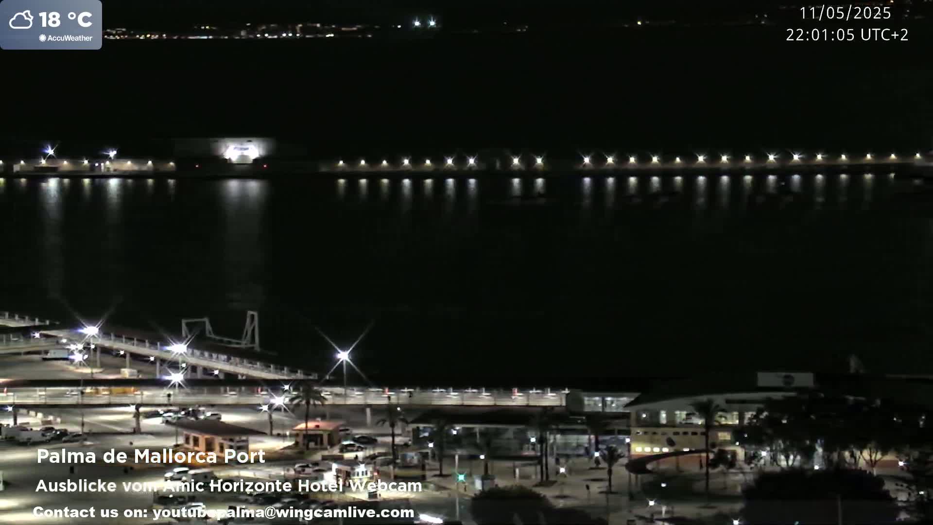 A nighttime view of a harbor with lights illuminating the water and a dock, under a clear sky with a temperature of 18 degrees Celsius.