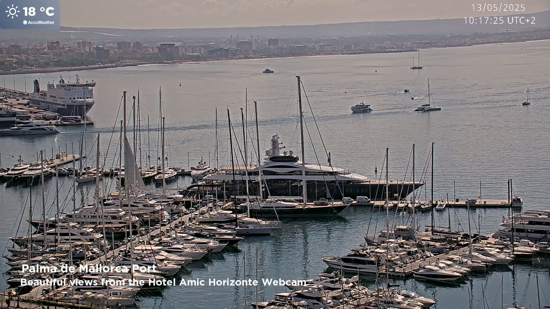 A large yacht is docked in a marina filled with numerous other boats, with a city and calm, sunny ocean visible in the background.