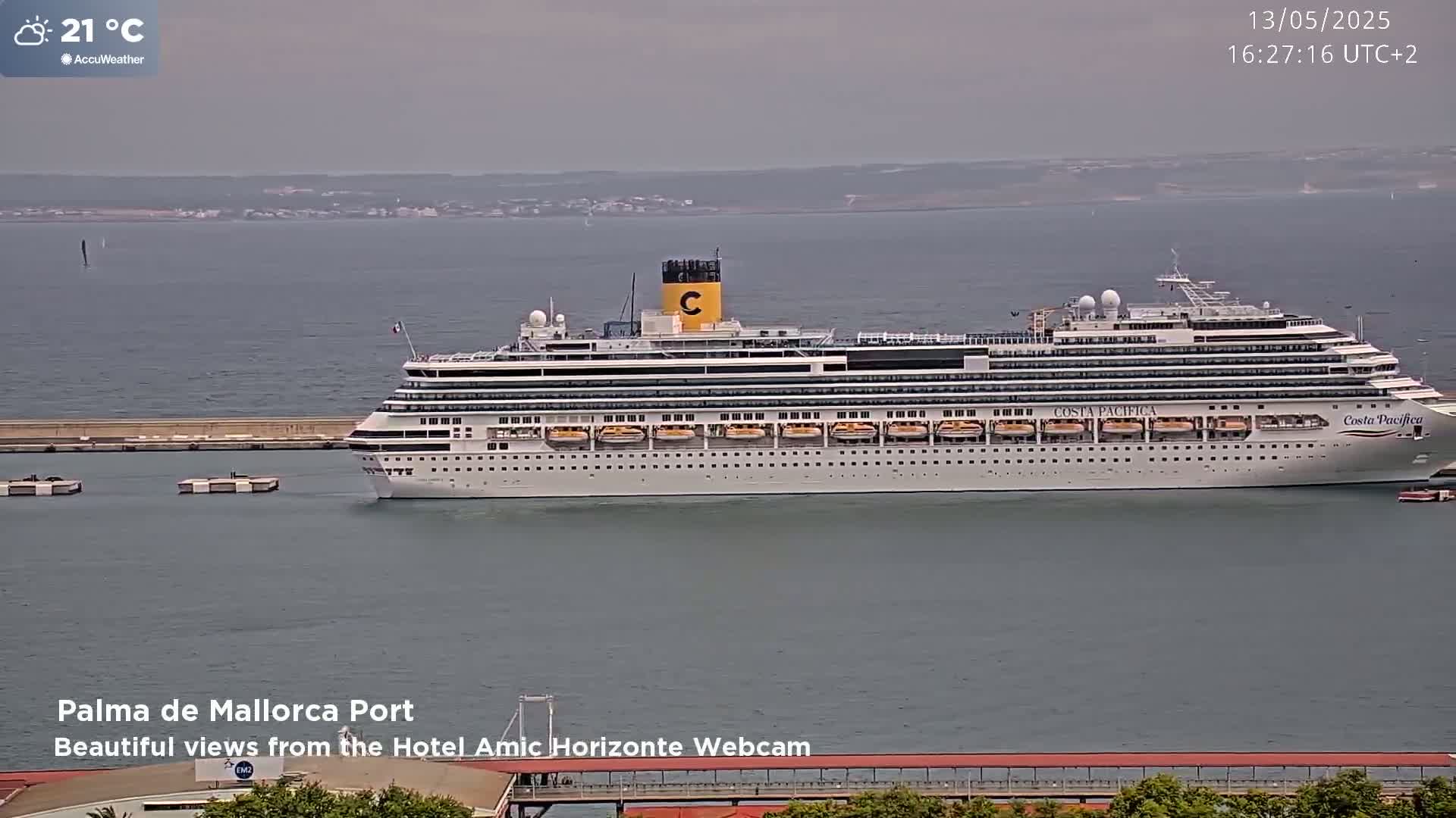 A large cruise ship is docked in a calm harbor on a slightly overcast day with a temperature of 21 degrees Celsius.