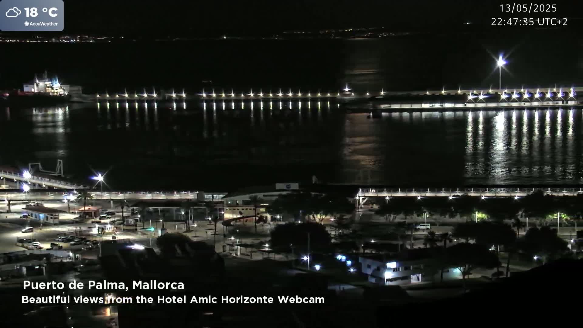 A nighttime view of a harbor with a ship, piers, and a bridge, under a partly cloudy sky with a temperature of 18 degrees Celsius.