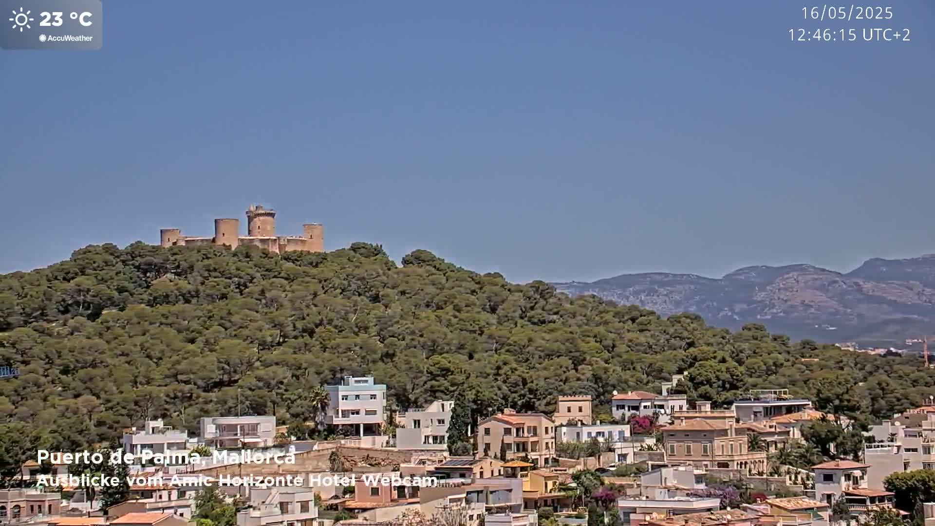 A stone castle sits atop a tree-covered hill overlooking a town and distant mountains under a clear blue sky.