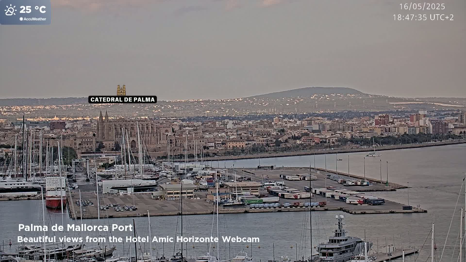 A harbor filled with sailboats and cargo containers is shown at dusk under a cloudy sky, with a city and a large cathedral in the background.
