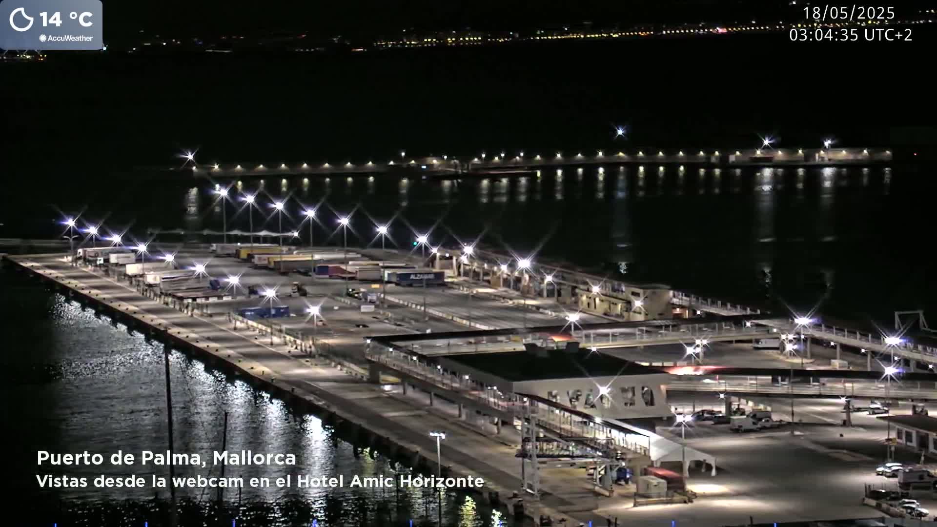A brightly lit port at night, with numerous vehicles parked on the docks and calm water reflecting the lights.