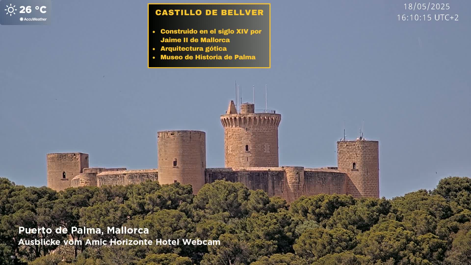 A stone castle with a central tower sits atop a hill covered in green trees under a clear blue sky.