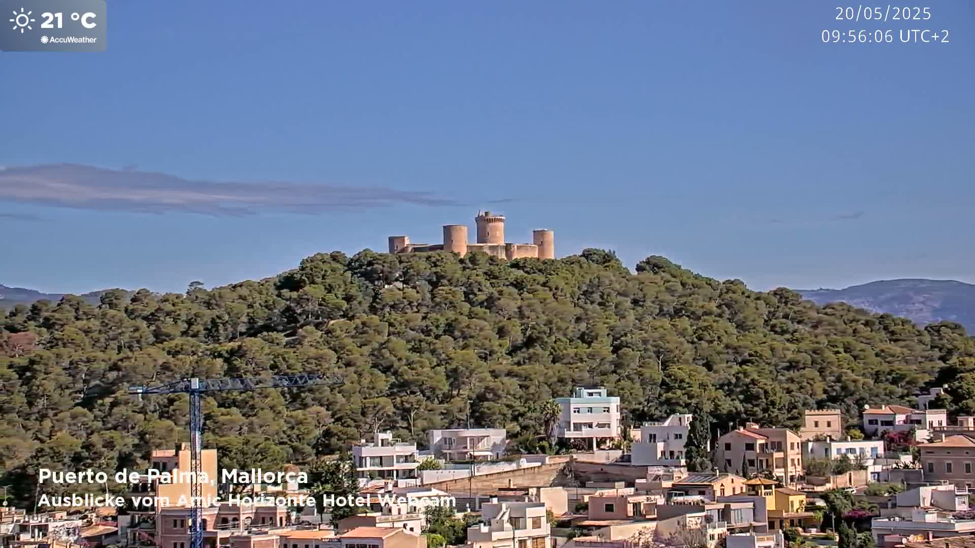 A stone castle sits atop a tree-covered hill overlooking a town of light-colored buildings under a clear blue sky.