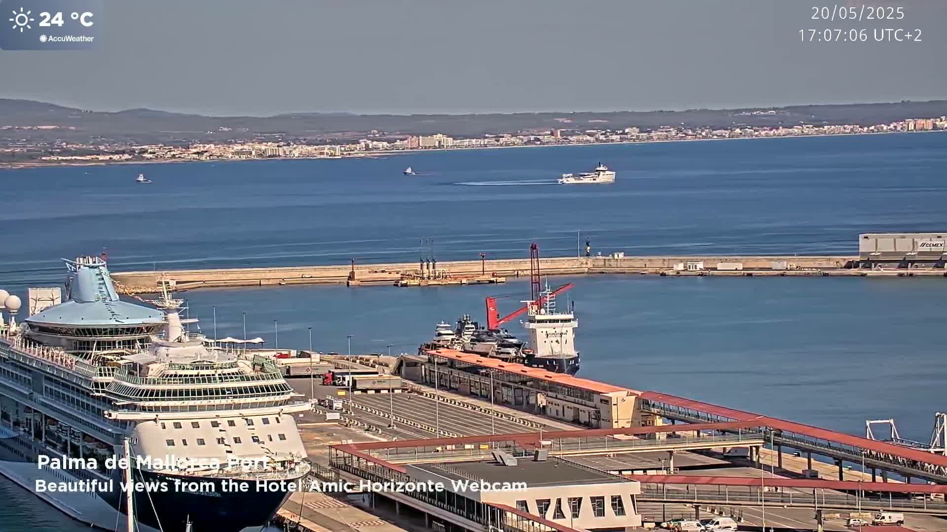 A cruise ship is docked at a port on a sunny day, with several other smaller vessels in the harbor and a city visible across the calm water in the distance.