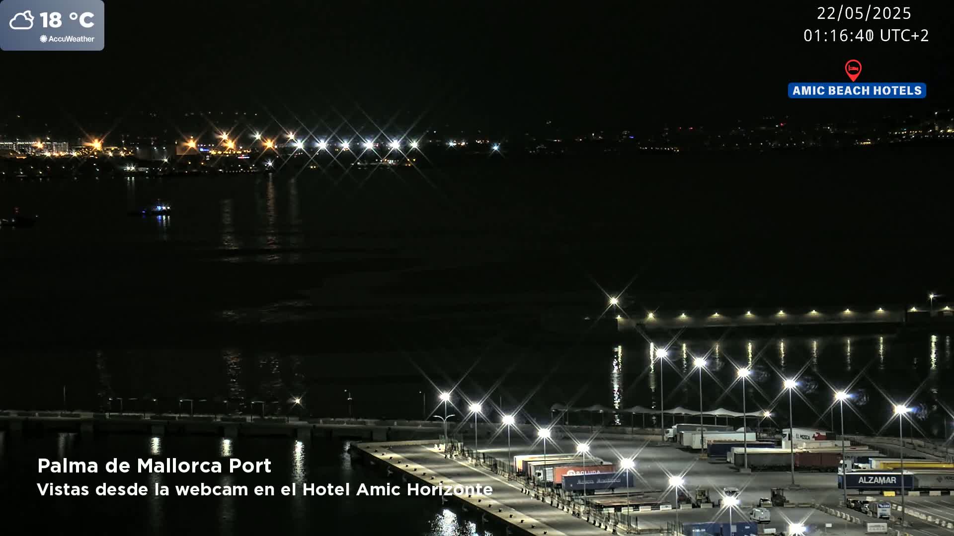 A nighttime view of a harbor with illuminated docks and a city skyline in the distance, under a clear sky.