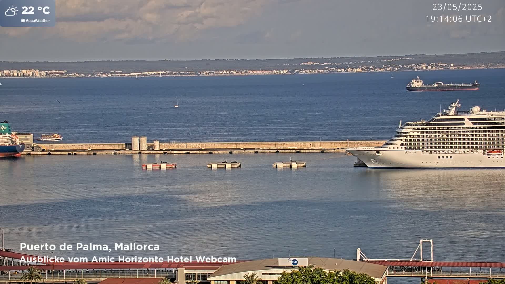 A large cruise ship is docked at a port, with other smaller vessels and a tanker in the distance, under partly cloudy skies and a temperature of 22 degrees Celsius.