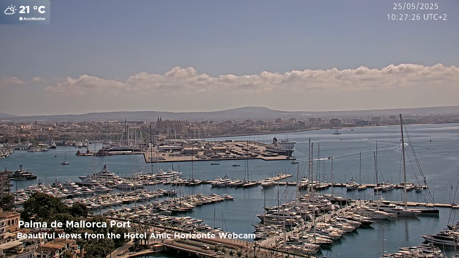 A harbor filled with numerous yachts and boats under a partly cloudy sky, with a city and distant mountains in the background, and a temperature of 21°C.