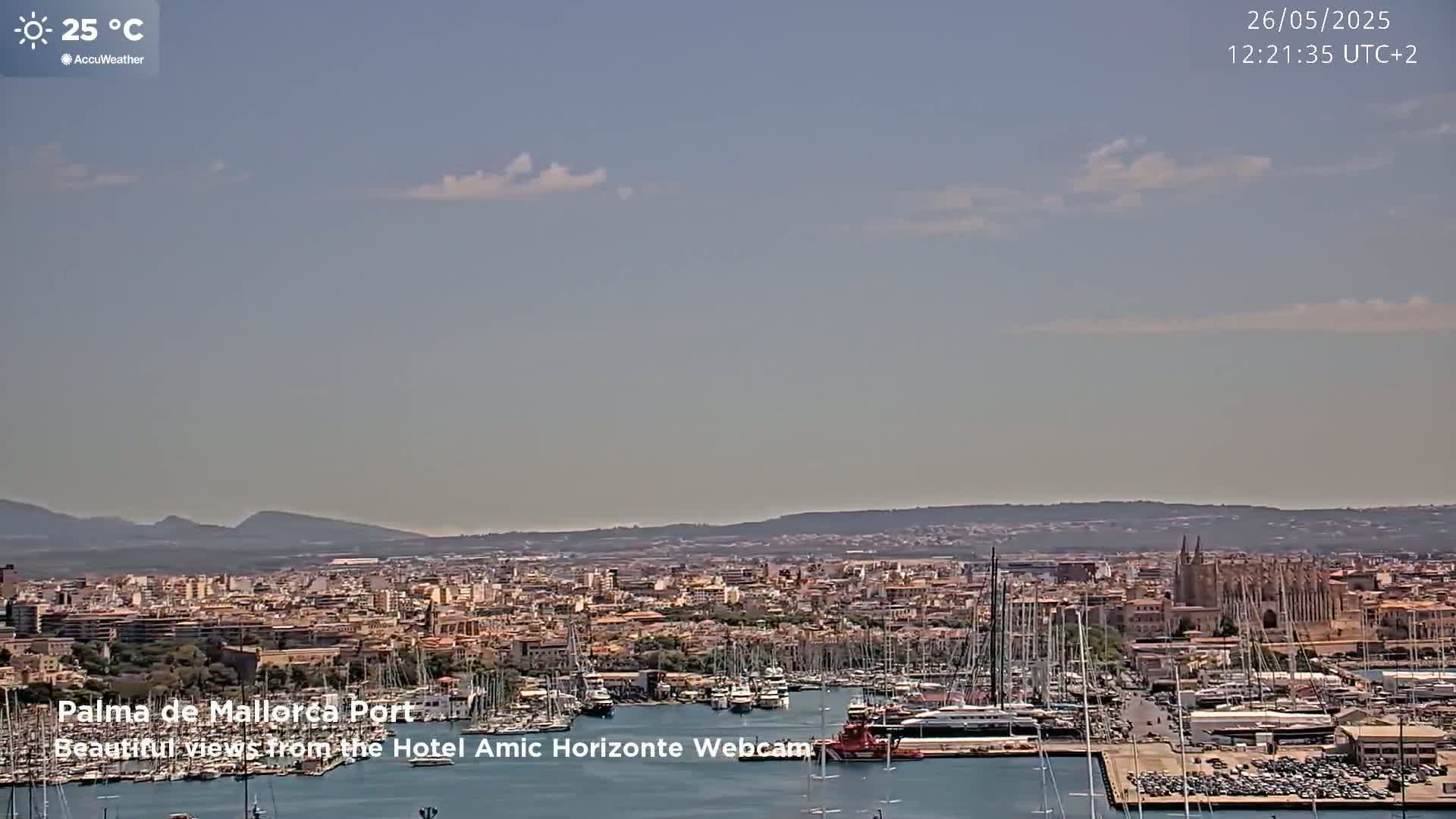 A partly cloudy, 25°C day overlooks a harbor filled with boats and a city sprawling against a mountain range in the distance.