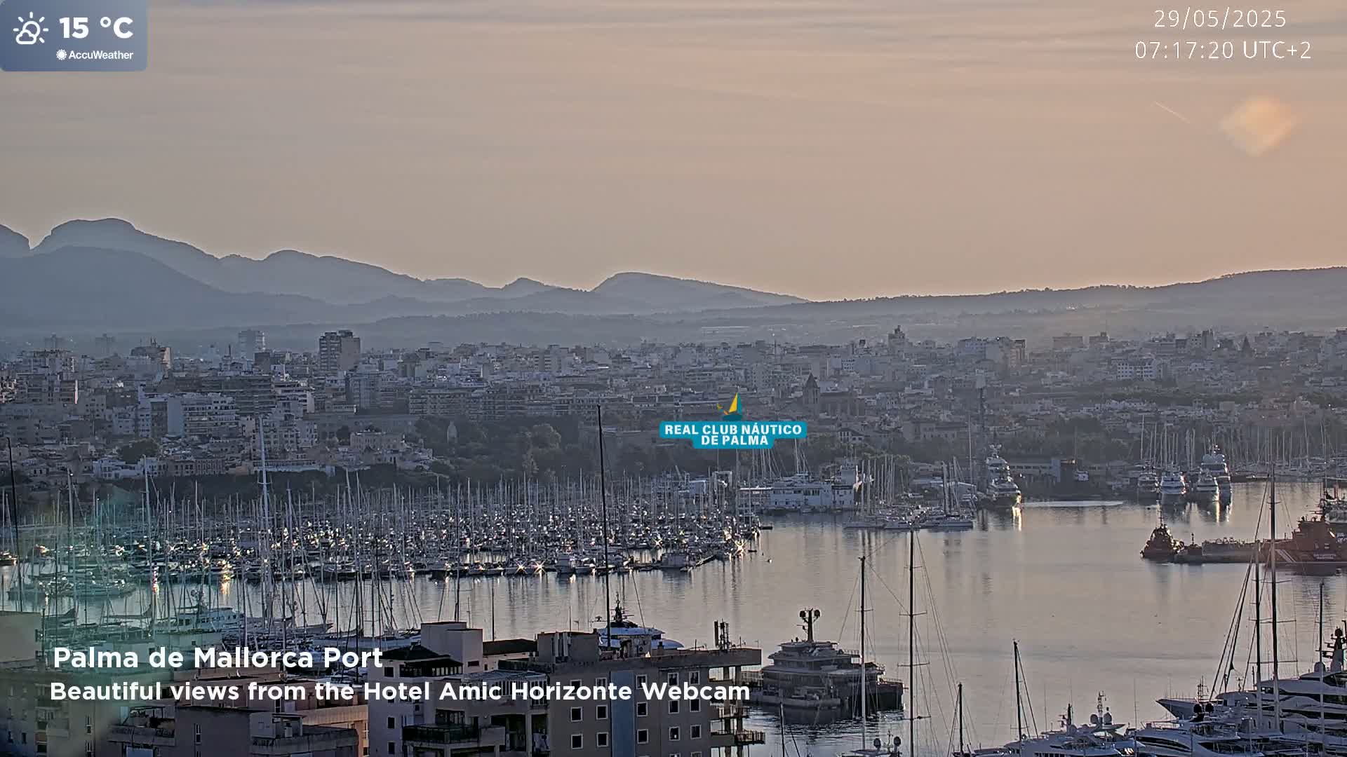 A harbor filled with numerous sailboats and yachts is situated in front of a city, all under a pale, hazy sky, with mountains visible in the distance.