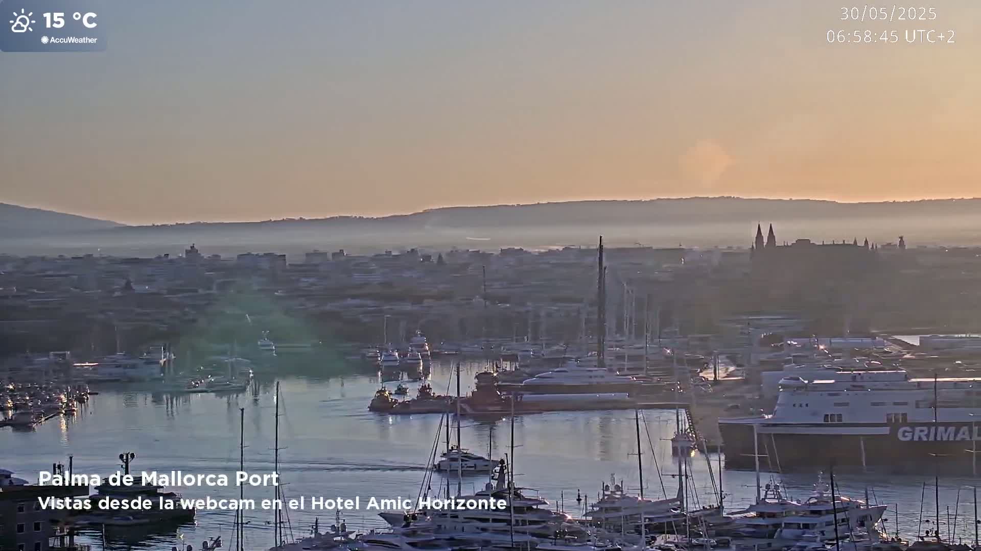 A hazy sunrise illuminates a harbor filled with numerous boats and yachts, with a city and distant mountains in the background.