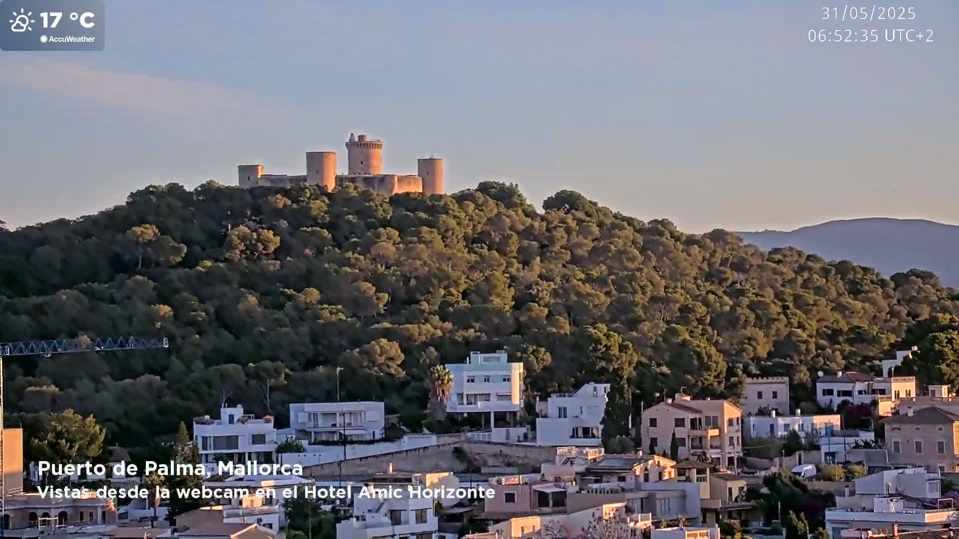 A stone castle sits atop a hill covered in lush green trees, overlooking a town of white and tan buildings under a clear, sunny sky.