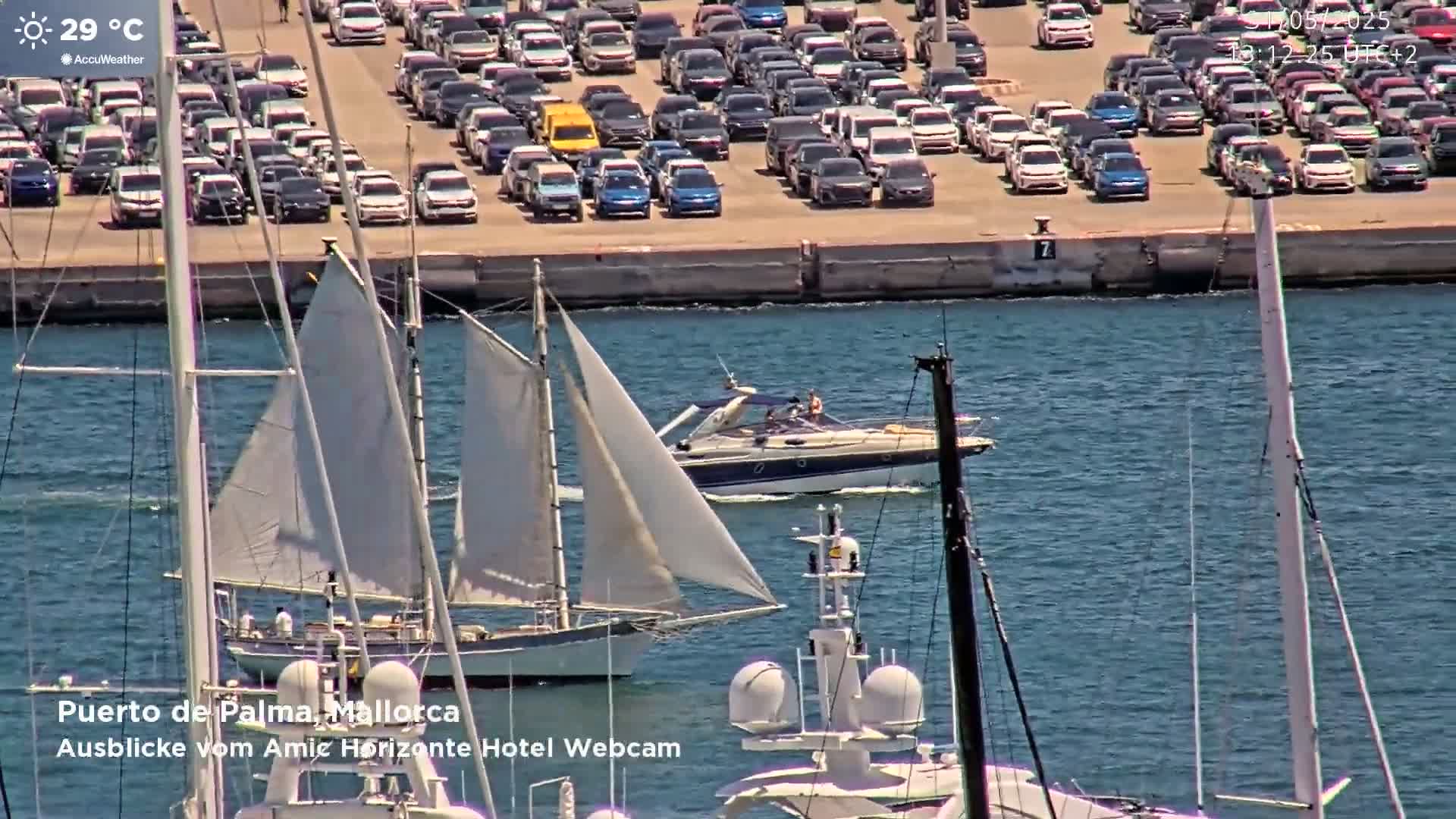 A sailboat and a motorboat are in a harbor next to a large parking lot filled with cars on a sunny day.