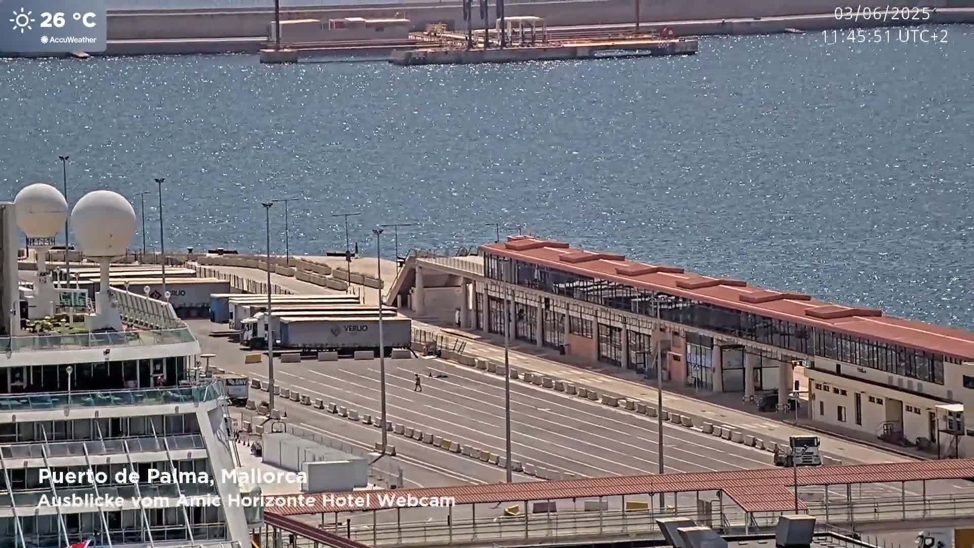 A cruise ship is docked at a port on a sunny day with calm, sparkling water in the background.