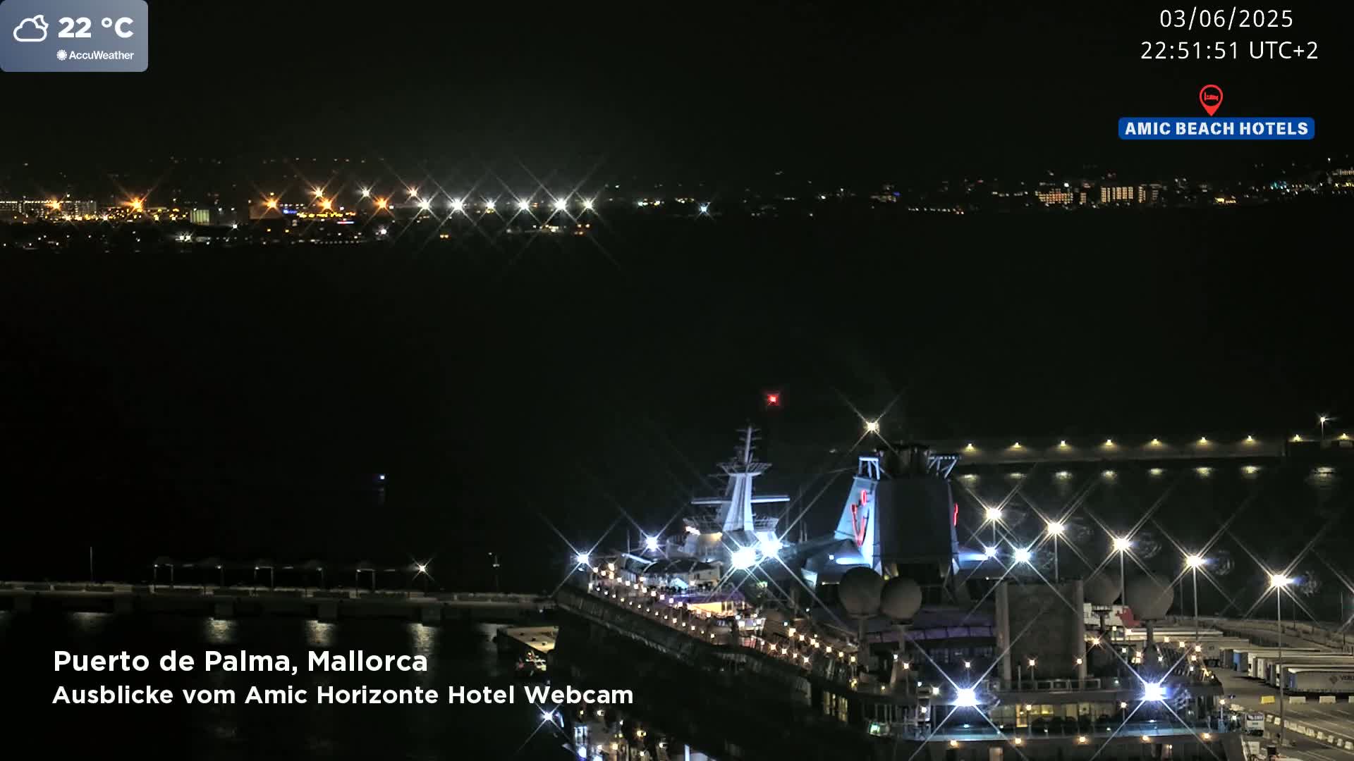 A brightly lit cruise ship is docked at a port at night, with a city's lights visible in the distance under a clear sky.