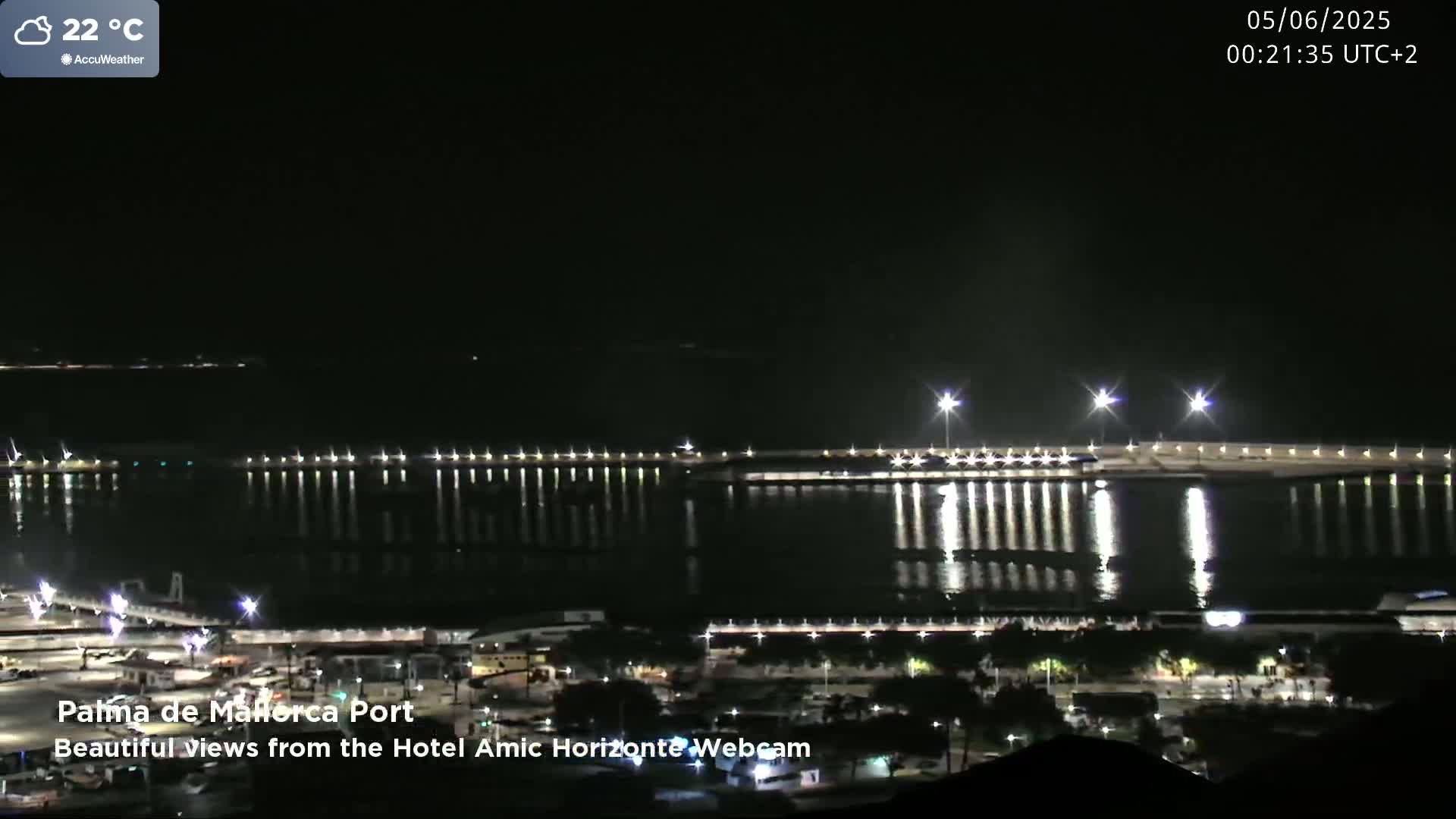 A nighttime view of a brightly lit harbor and city, reflecting in calm water under a clear sky.