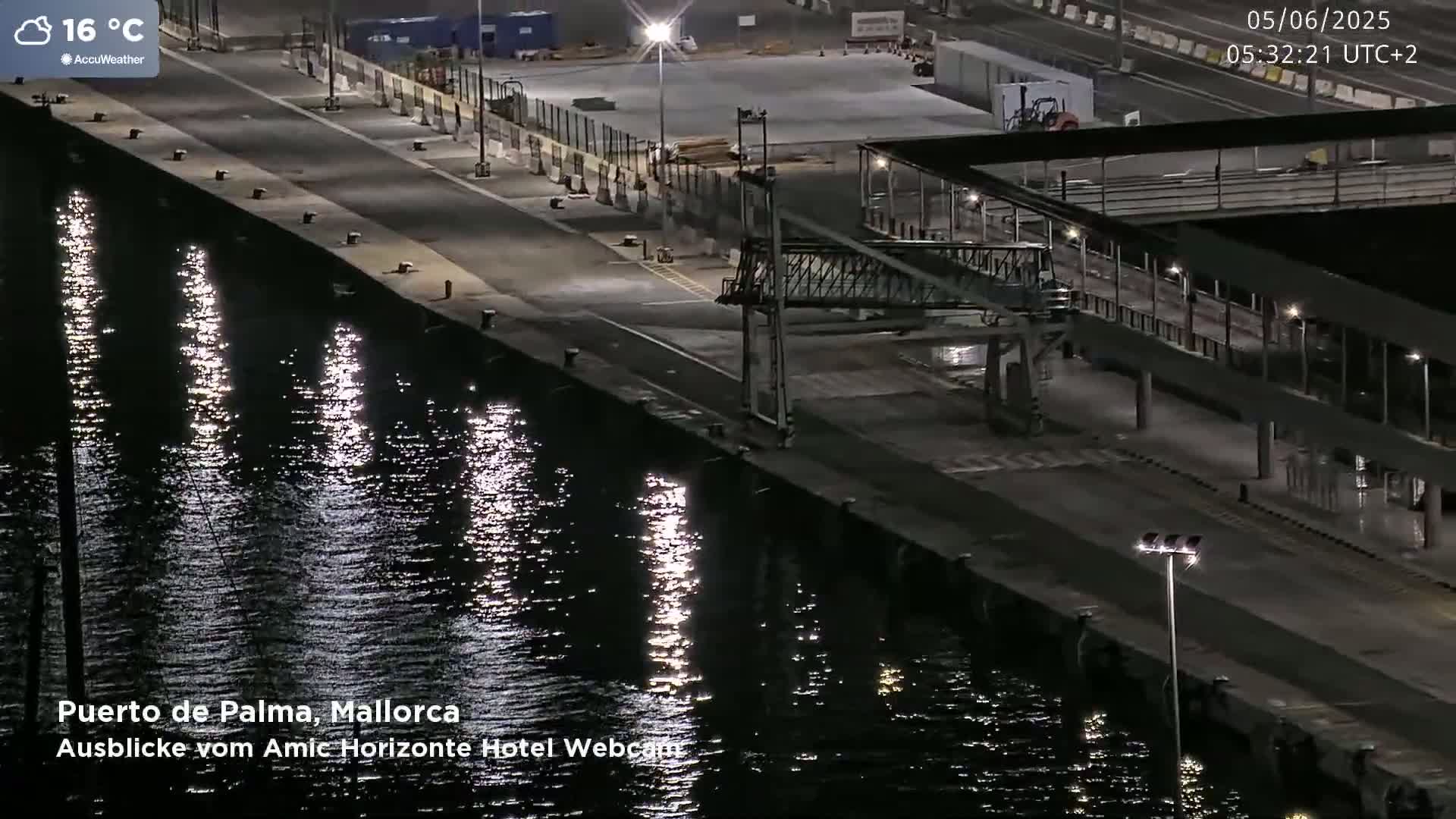 A nighttime view of a harbor with calm water reflecting lights, a dock with a walkway and building, and a mostly empty parking area.