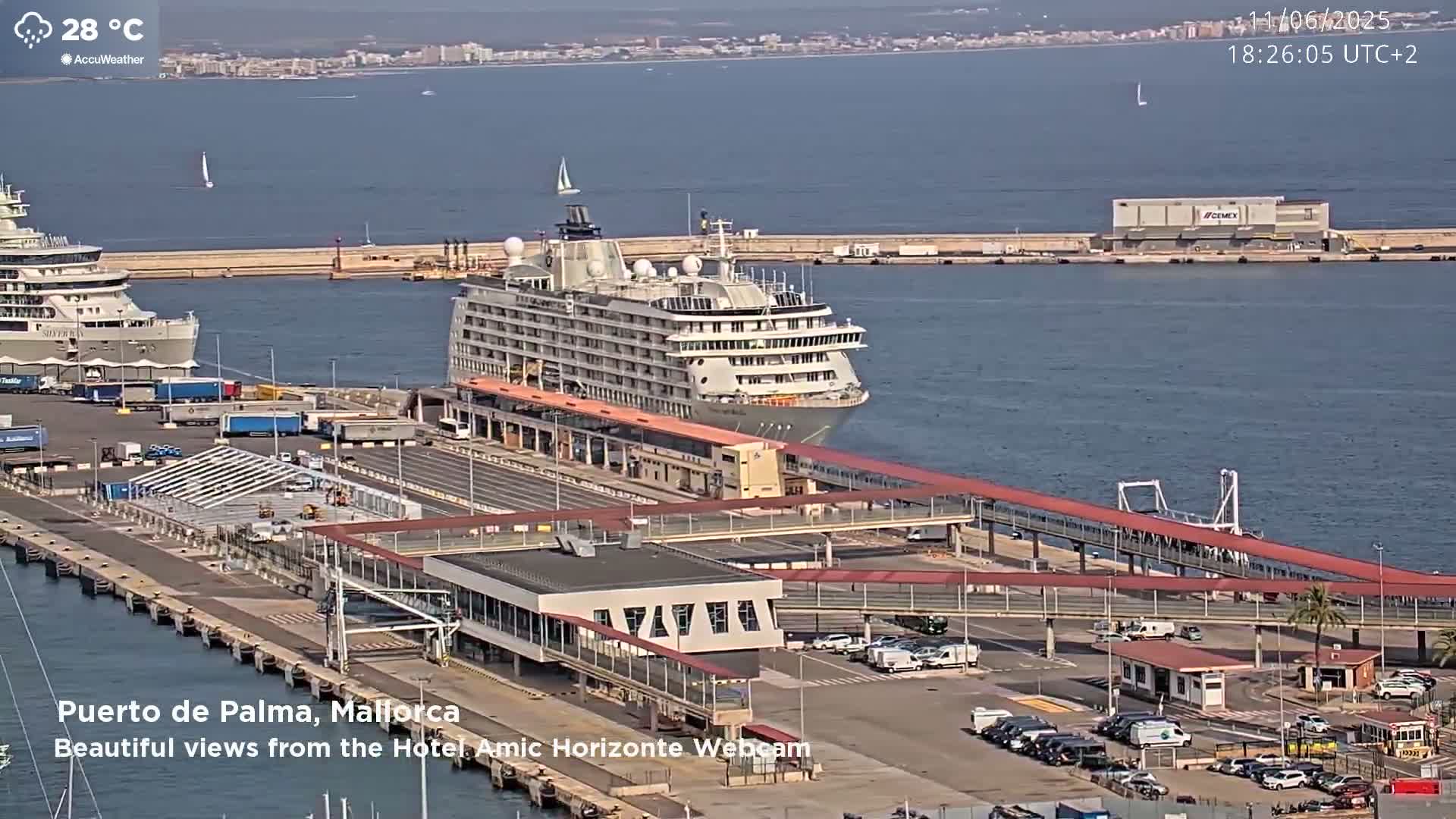 A large cruise ship is docked at a port on a sunny day, with other ships and vehicles visible in the harbor.
