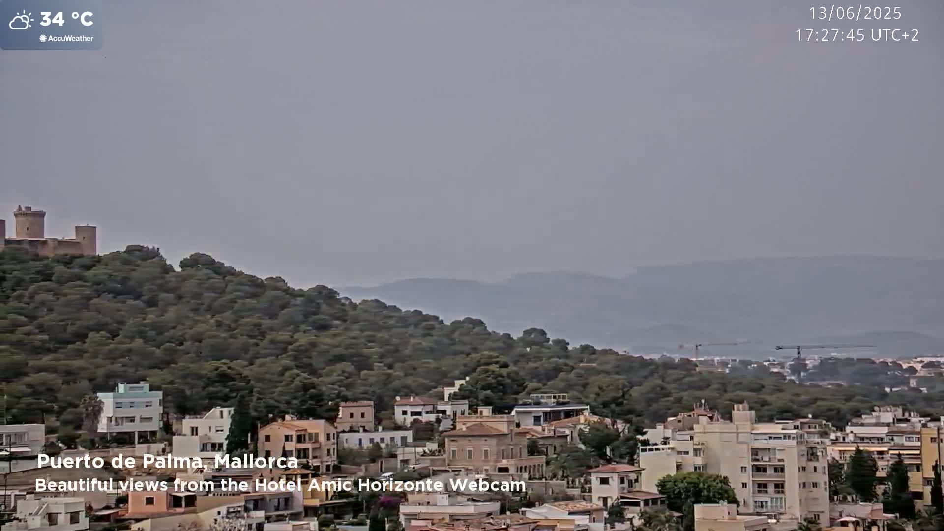 A hazy, 34°C day shows a hilltop castle overlooking a town nestled amongst green hills and distant mountains.