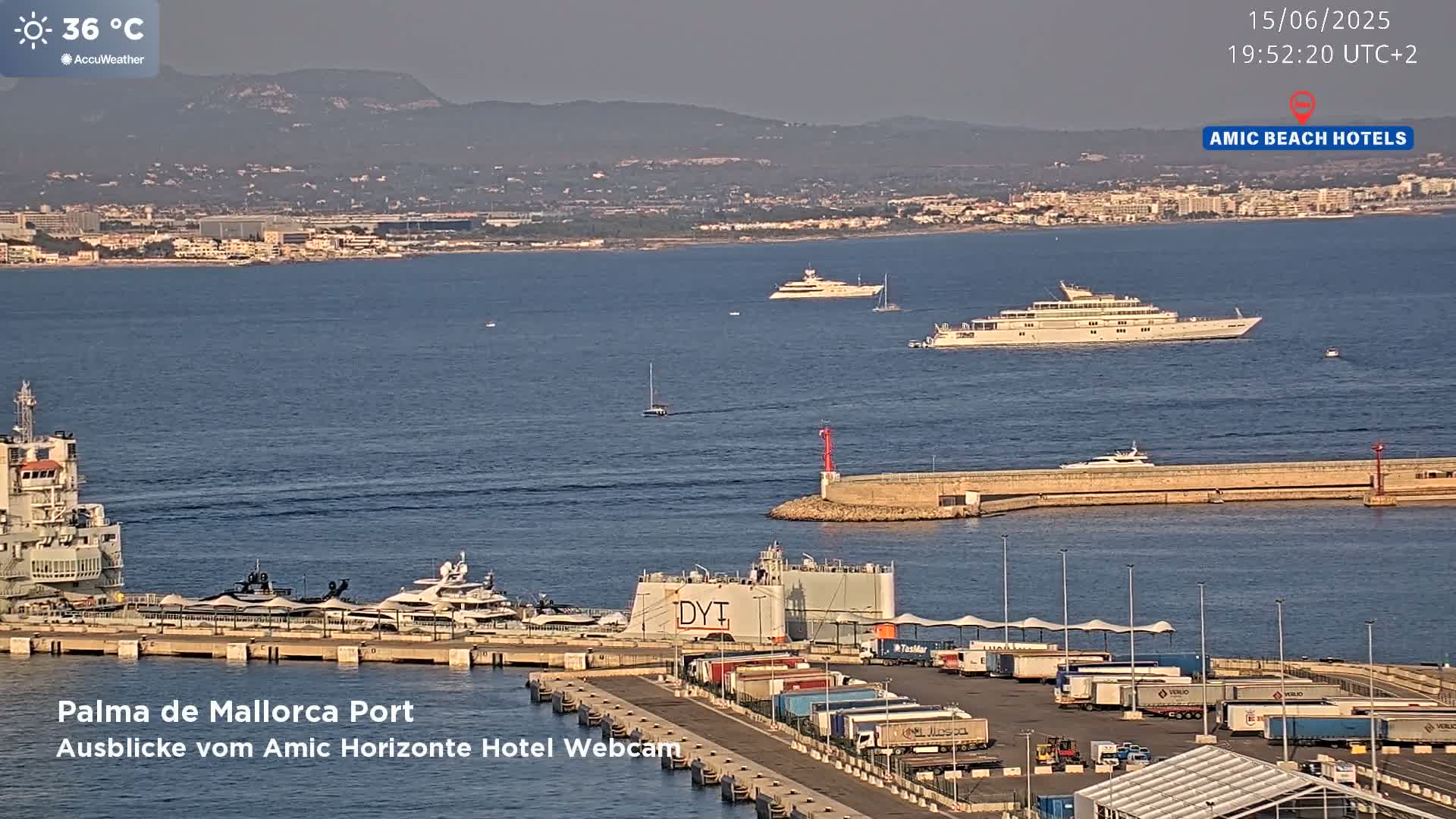 A harbor filled with several yachts and cargo trucks under a clear, sunny sky with a temperature of 36 degrees Celsius.