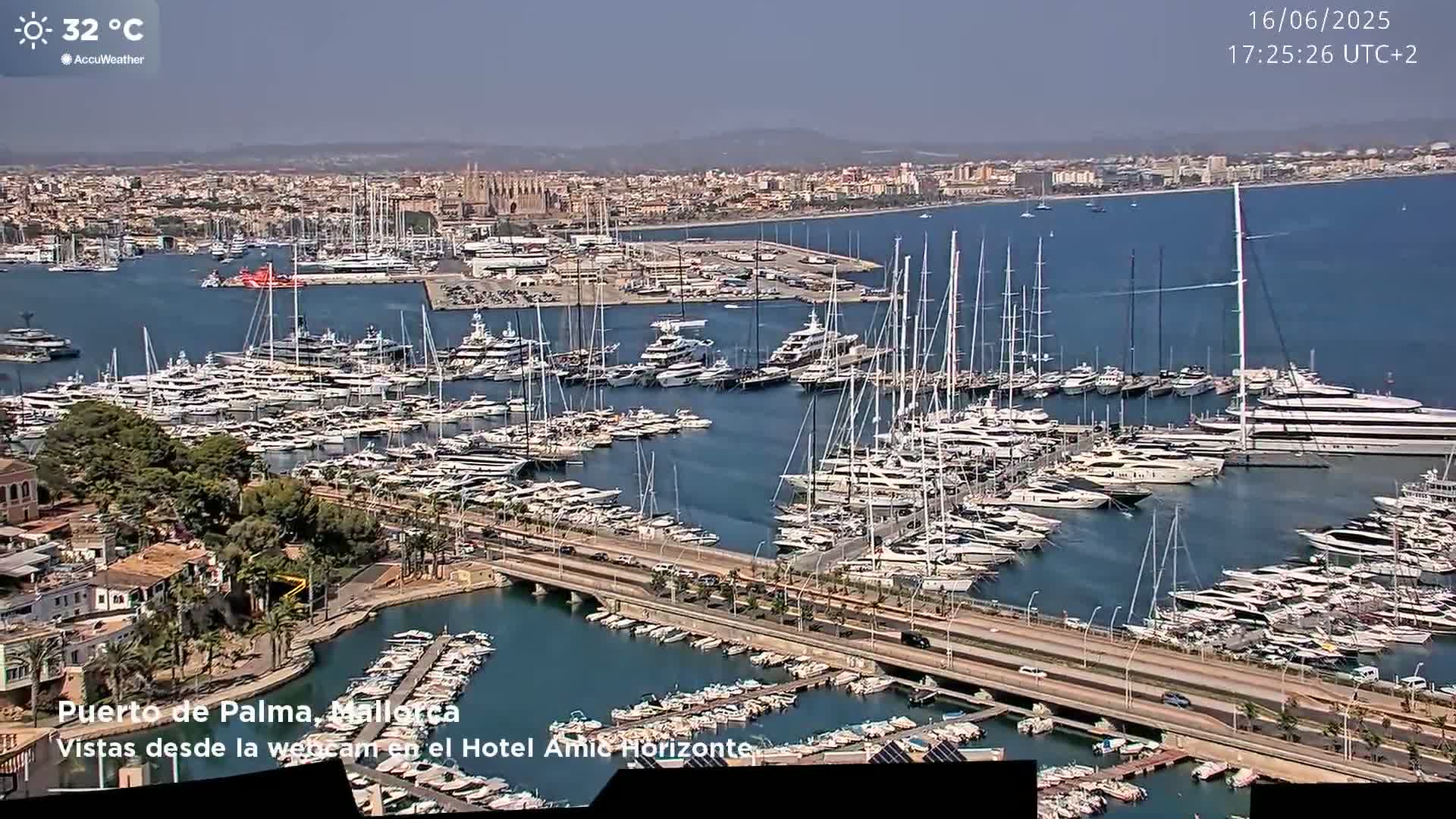 A harbor filled with numerous yachts and sailboats on a sunny day with a temperature of 32°C, overlooking a city.