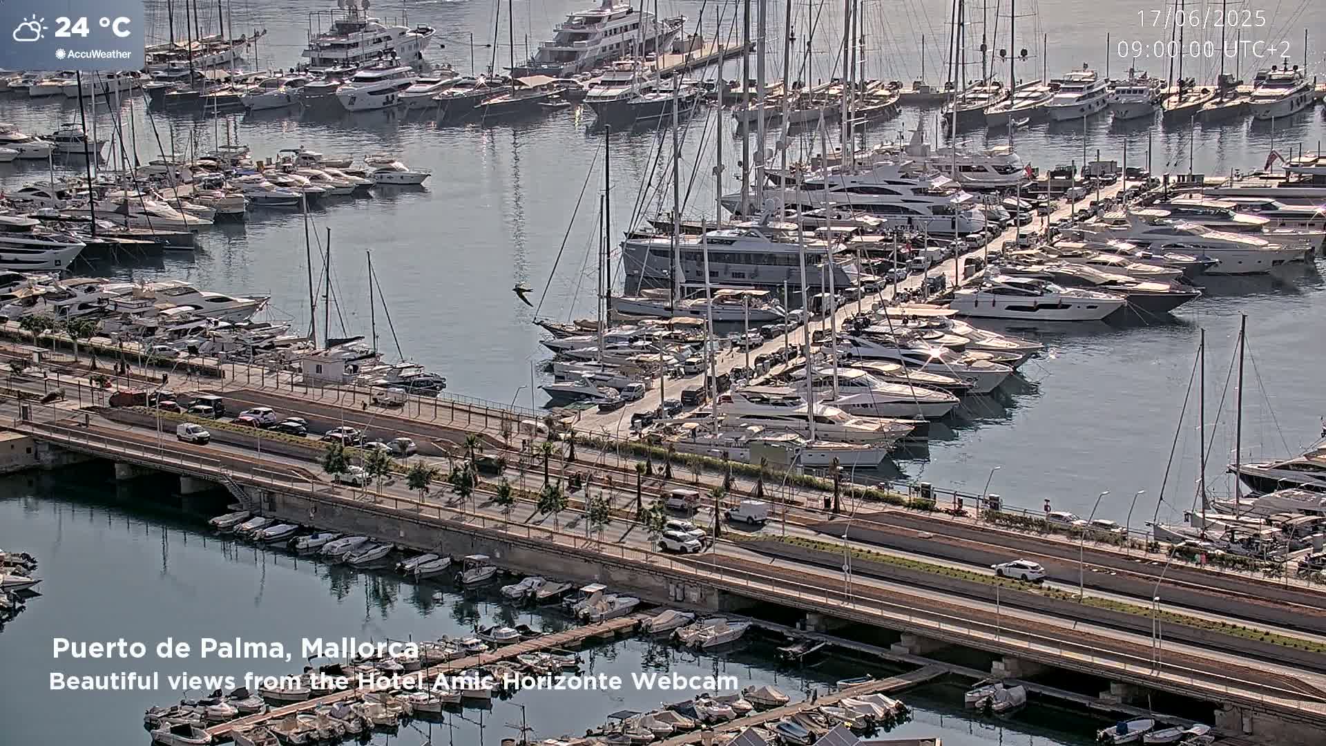 A marina filled with numerous yachts and sailboats on a sunny day, with a road and bridge visible alongside.