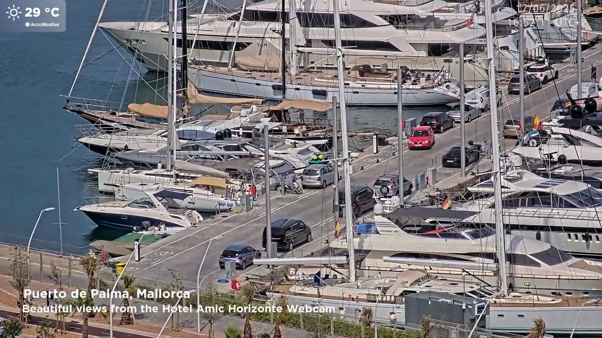 A marina filled with yachts and sailboats on a sunny day with a temperature of 29 degrees Celsius, with cars parked along a roadway next to the water.