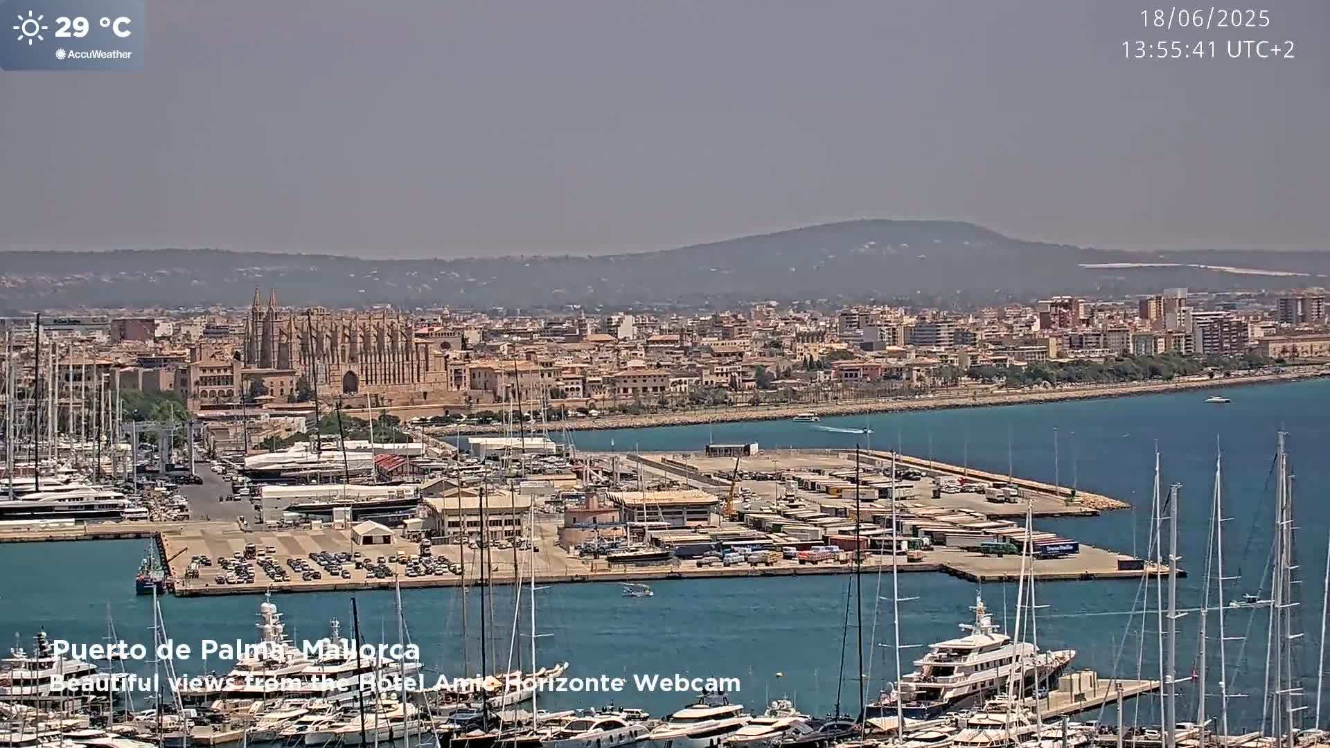A harbor filled with numerous boats and yachts is situated before a city featuring a large cathedral, all under a hazy, sunny sky with a temperature of 29 degrees Celsius.