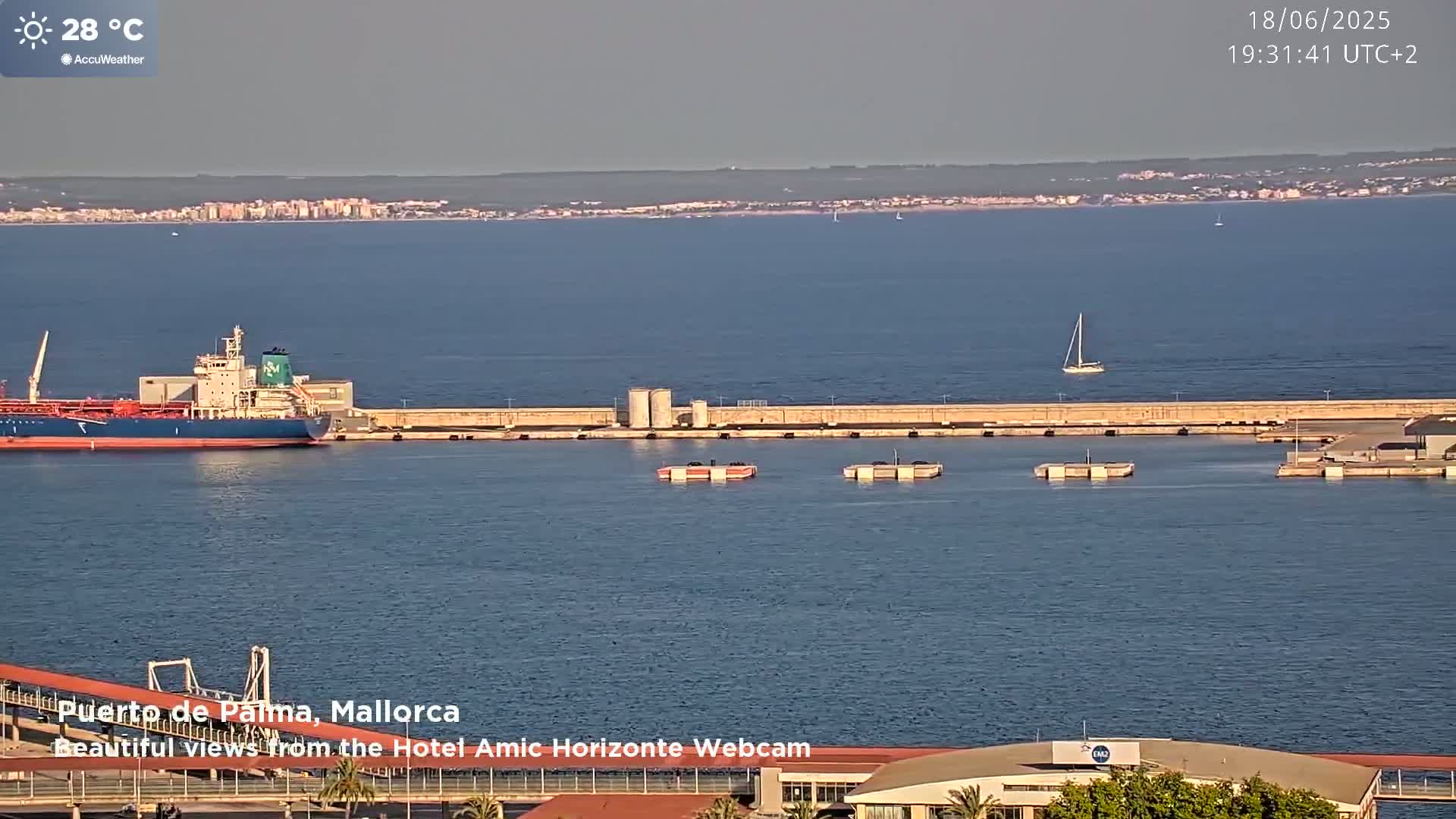 A large tanker sits in a calm harbor on a sunny day, with a distant coastline and a sailboat visible in the bay.