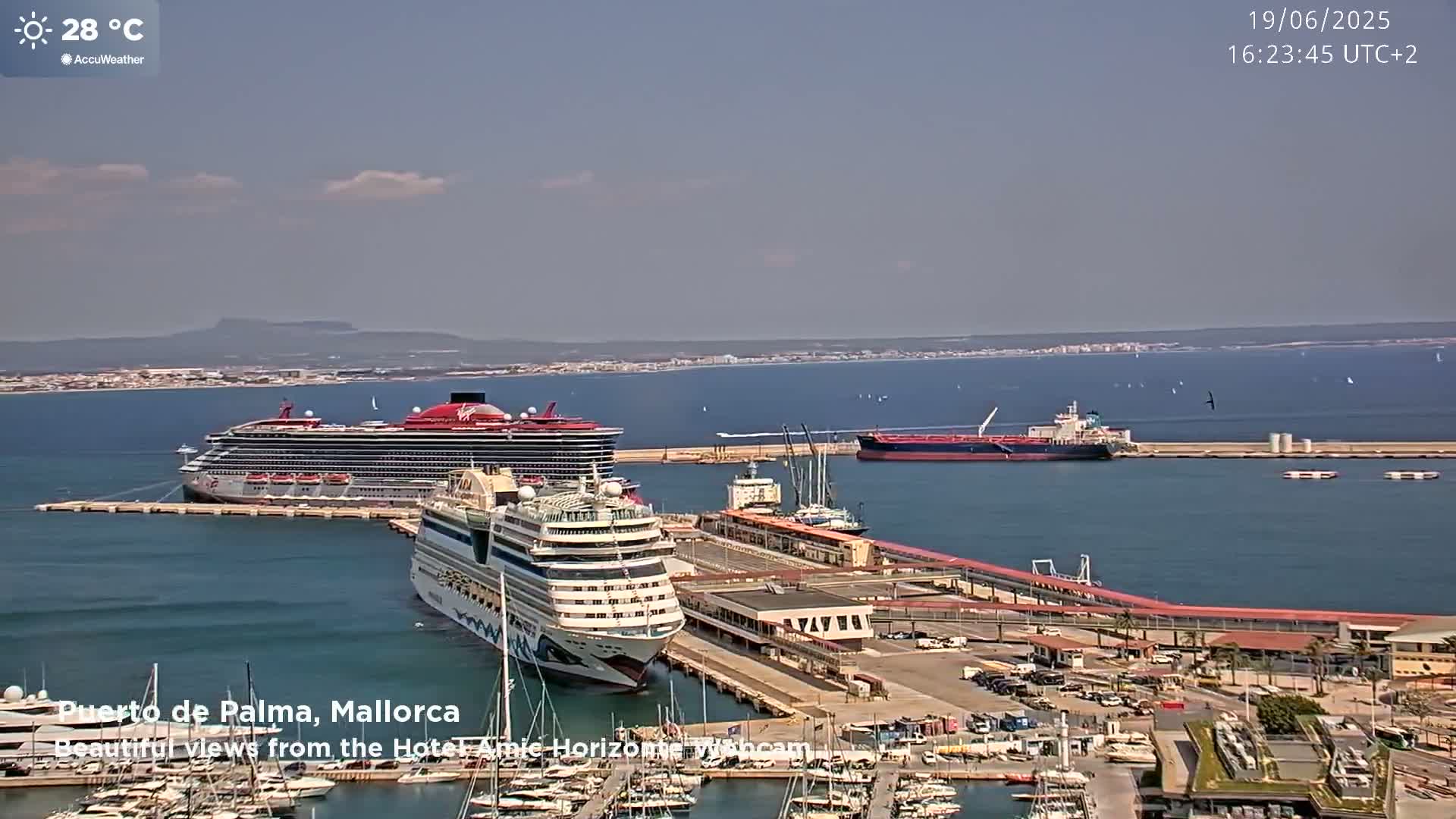 Two cruise ships are docked at a port on a sunny day, with a tanker ship and numerous smaller boats in the harbor.