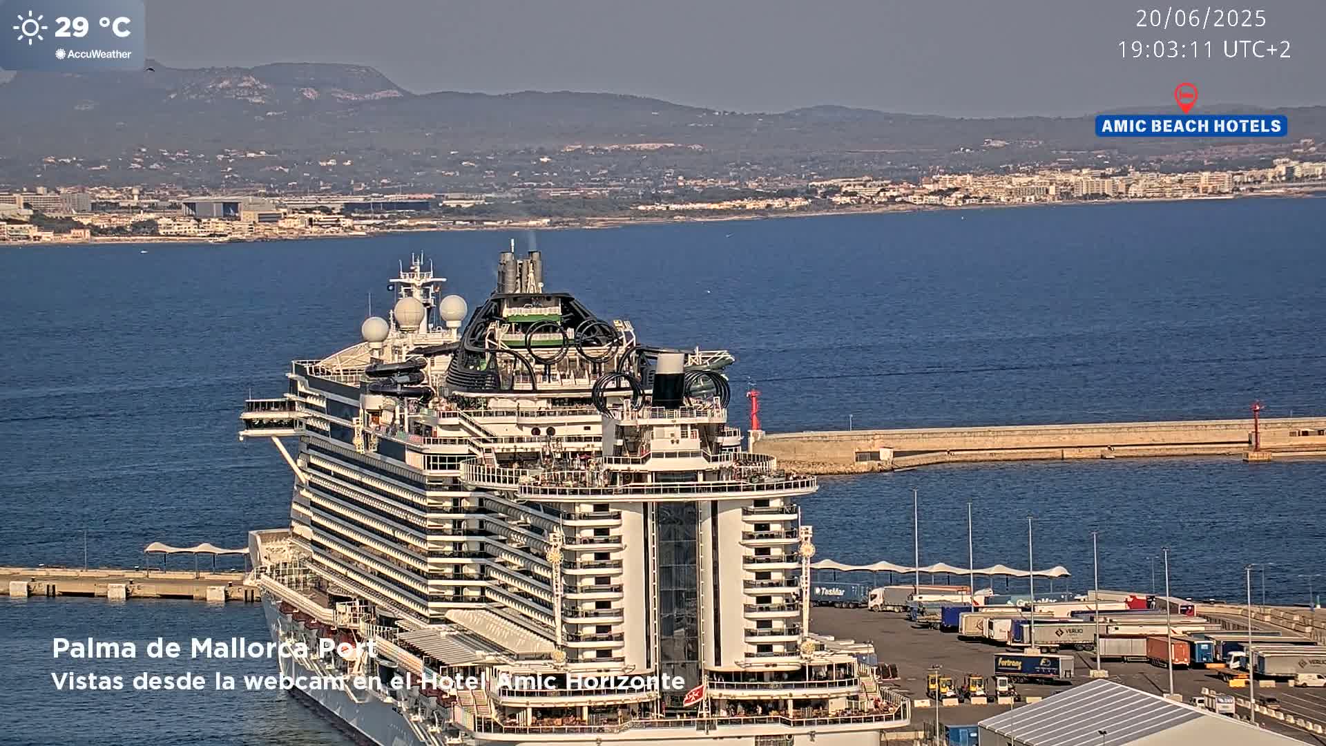 A large cruise ship is docked at a port on a sunny day, with a city and mountains visible in the distance.