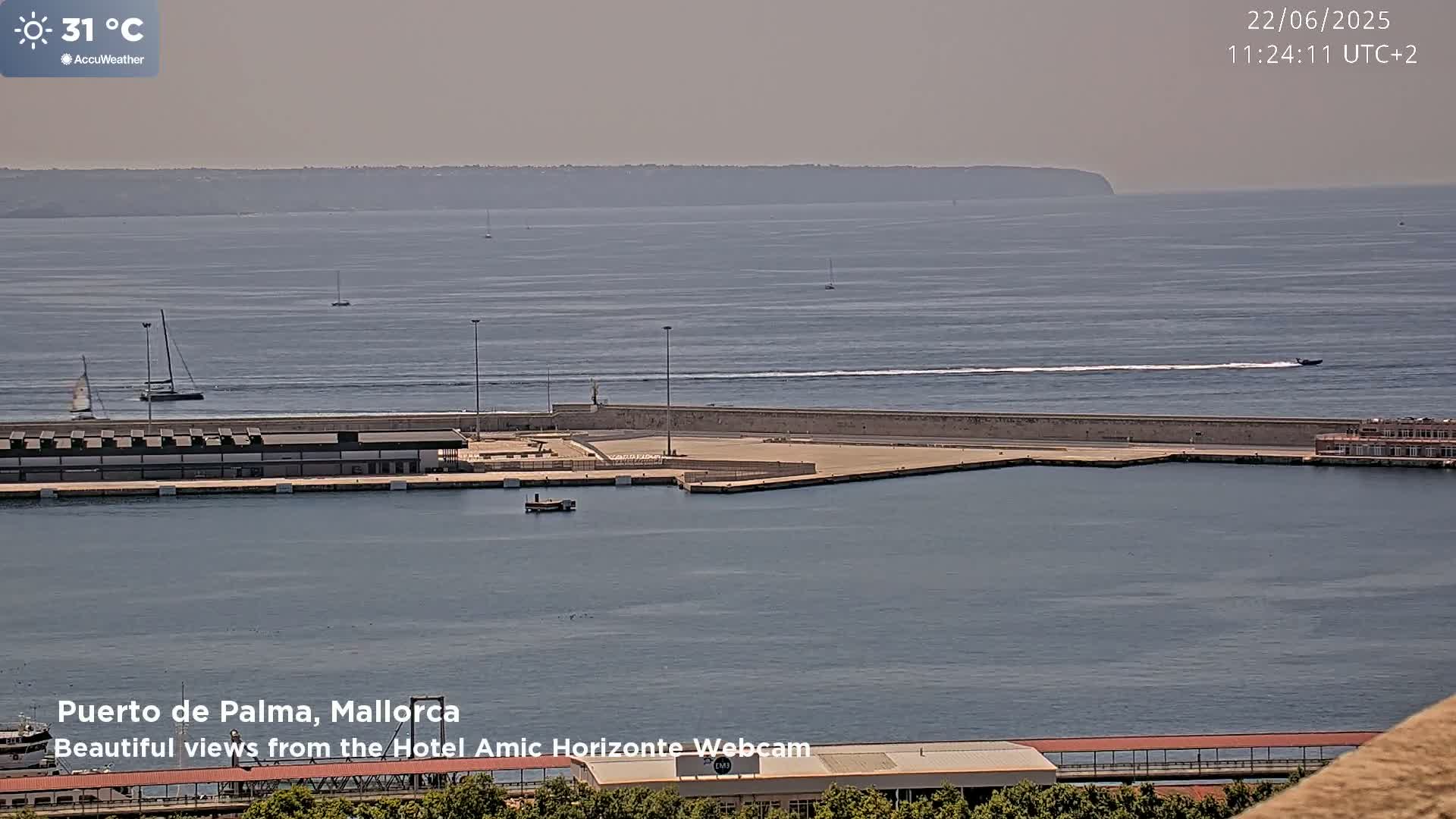 A harbor with several boats, including sailboats and a speedboat, is shown on a hazy, sunny day with a distant landmass visible across calm water.
