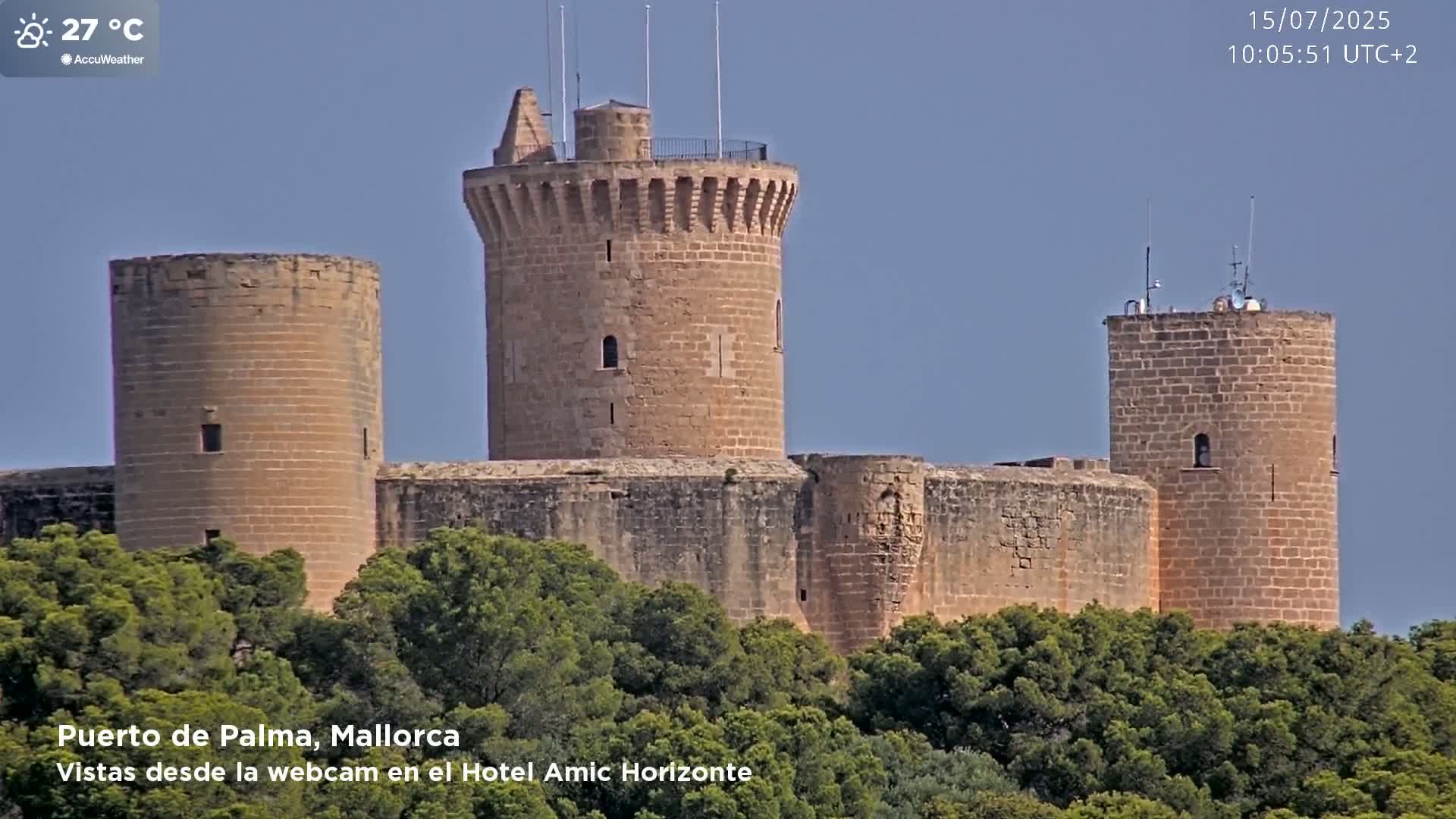 A stone castle with multiple towers is partially obscured by green trees under a clear, sunny sky.