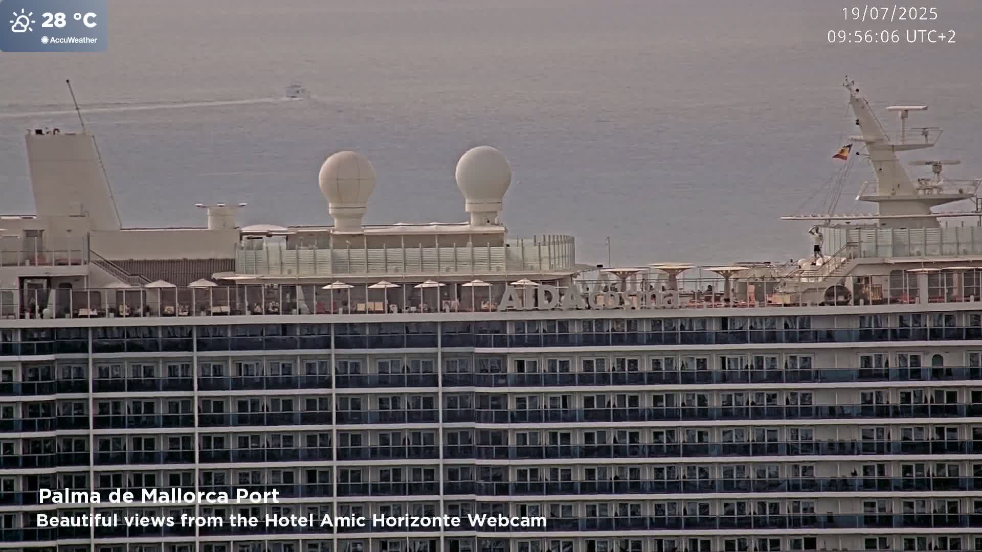 A large cruise ship with multiple decks of balconies is docked at a port on a hazy day with calm water visible in the background.
