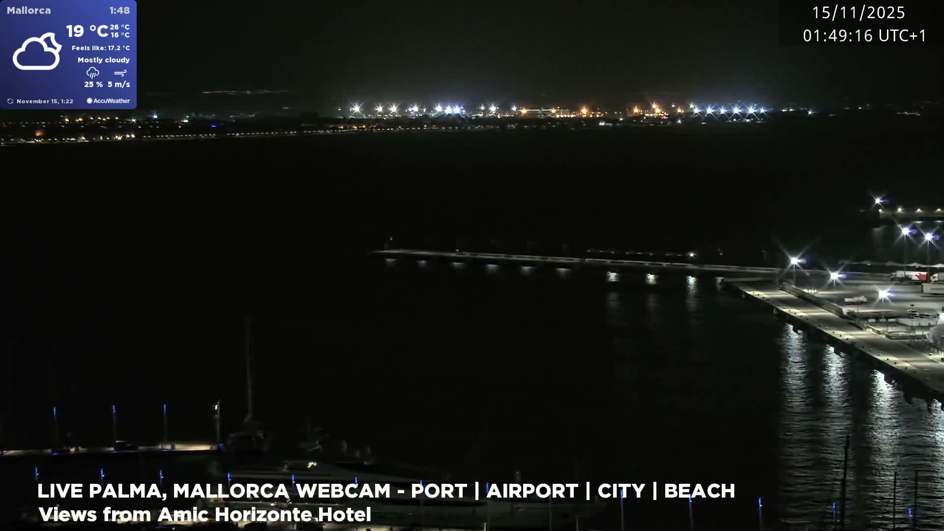 A wide nighttime scene overlooks a dark body of water, featuring a brightly lit pier with sparse activity in the foreground, and numerous lights illuminating a distant city or port along the horizon.