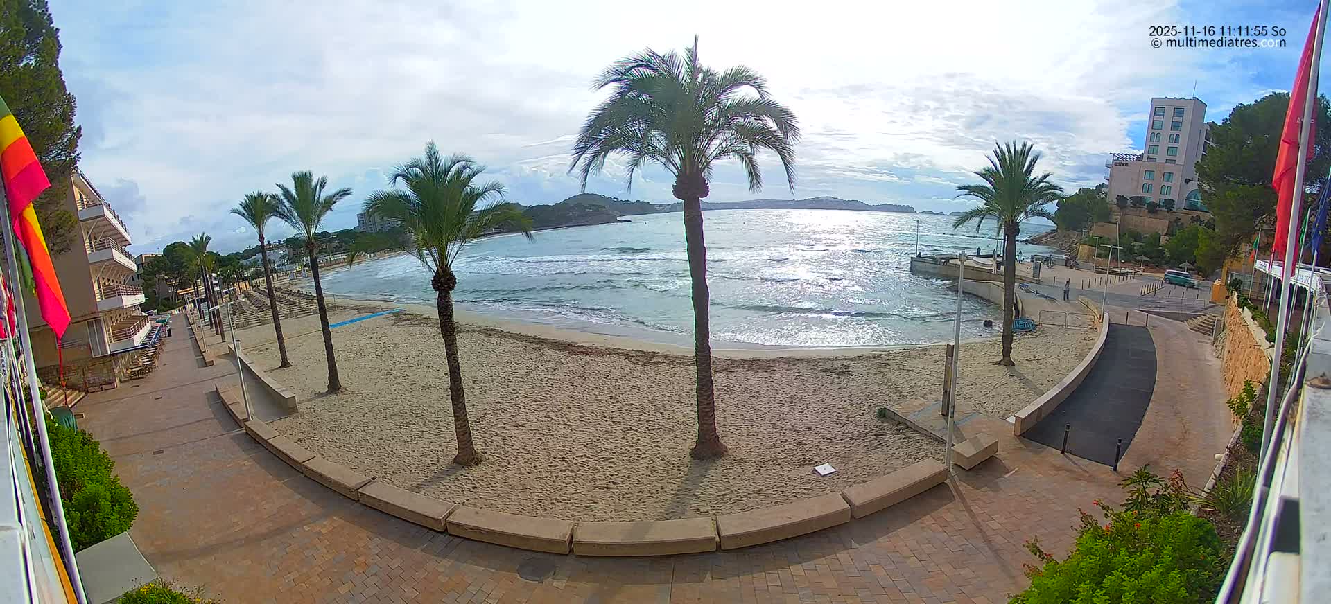 A wide-angle view captures a sandy beach with palm trees and a small pier, where gentle waves meet the shore under a bright, partly cloudy sky, with resort buildings lining the promenade.