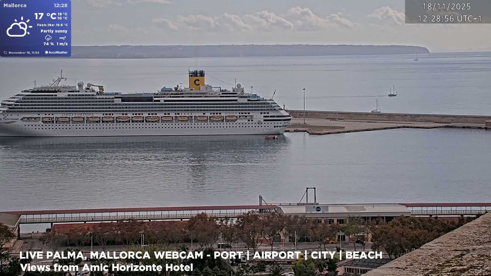 A wide-angle view captures a sandy beach with palm trees and a small pier, where gentle waves meet the shore under a bright, partly cloudy sky, with resort buildings lining the promenade.