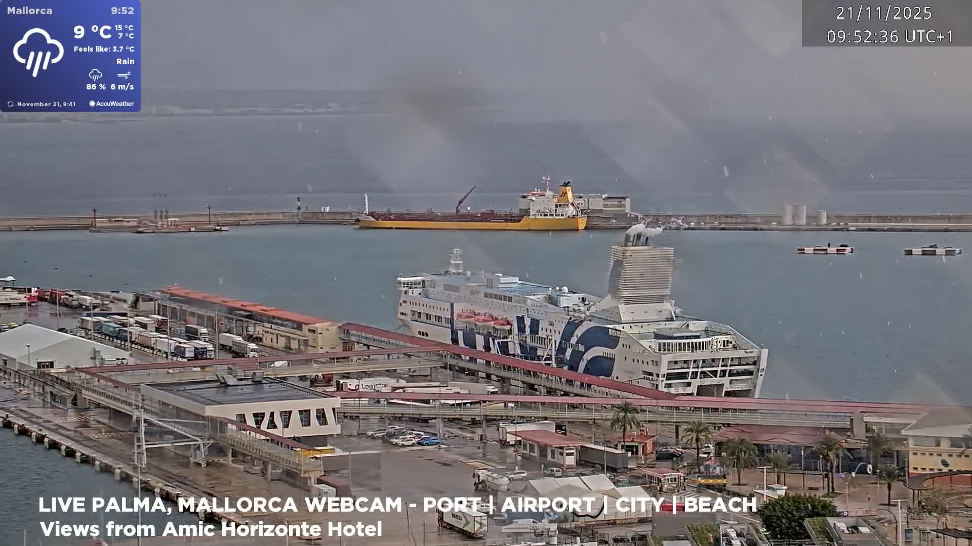 A rainy day unfolds at a busy port, showcasing a large white and blue ferry docked at a pier amidst various port buildings and parked vehicles, with a yellow cargo ship visible further out in the grey harbor under an overcast sky.