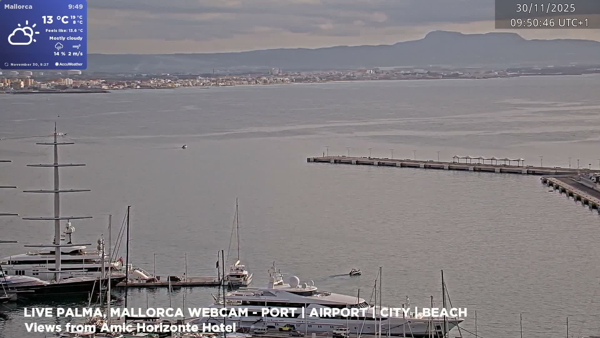 A cloudy day overlooks a harbor filled with various docked boats and yachts, with a long pier extending into the water and a distant coastal city backed by mountains under an overcast sky.